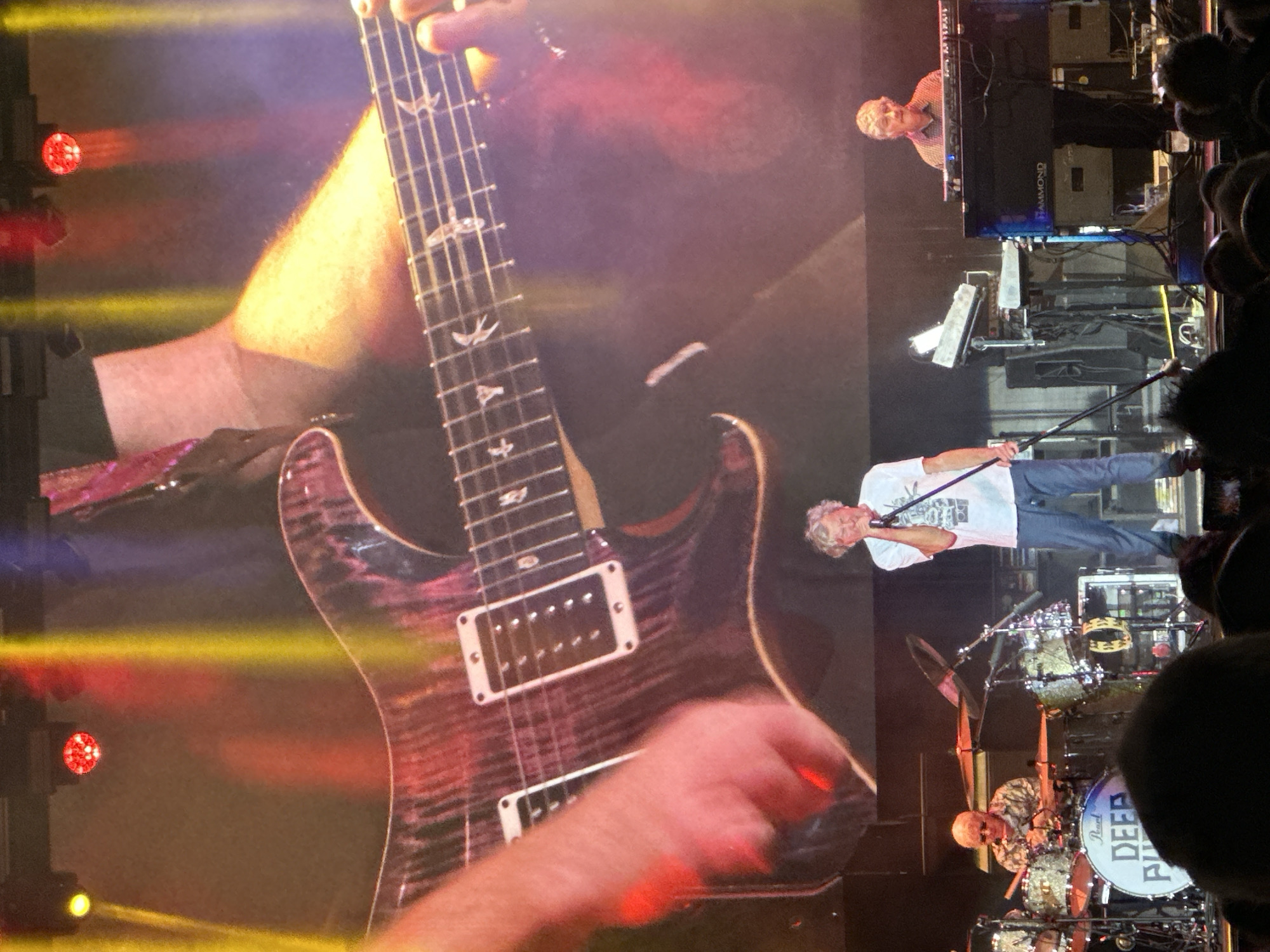 A concert is taking place on a stage in Jerez De La Frontera, Spain. A large screen behind the performers displays a close-up of a person's hand playing an electric guitar, with colorful stage lights creating streaks of red, yellow, and blue. In the foreground, on the stage, three musicians are visible. To the left, a drummer with glasses is seated behind a drum kit, which features a large bass drum with the "DEEP PURPLE" logo. In the center, a man with gray hair is singing into a microphone, wearing a white t-shirt and blue jeans. To the right, another man with gray hair is playing a keyboard, with the "HAMMOND" logo visible on the instrument. The audience is visible in the lower portion of the image, with many heads and shoulders in silhouette.
