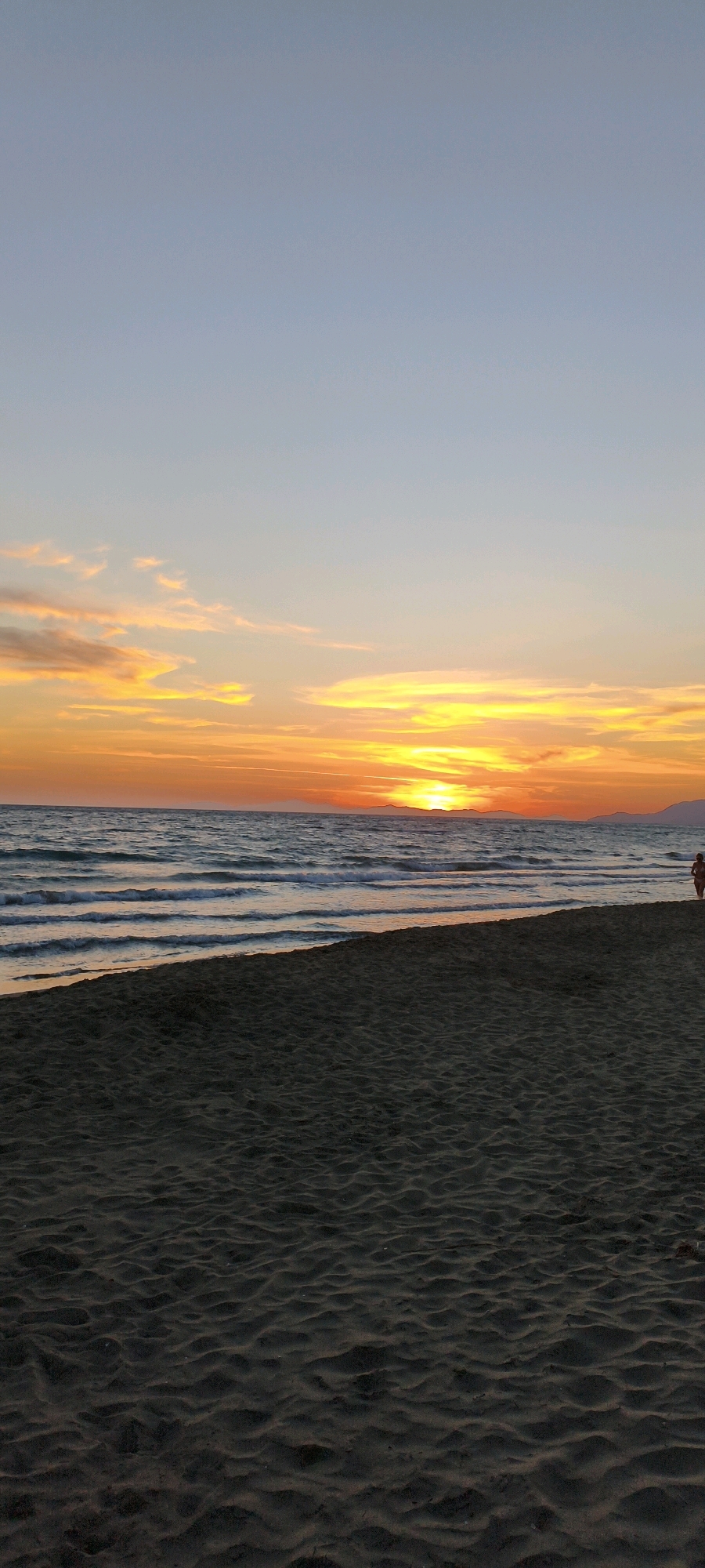 The image shows a beach at sunset. The sky is a gradient of blue, fading into orange and yellow near the horizon. Wispy clouds are scattered across the sky, illuminated by the setting sun. The ocean has gentle waves and reflects the colors of the sunset. The foreground is a sandy beach with ripples from the receding tide. In the distance, a person is walking along the shoreline. The scene appears to be the coast of the Maremma region in the province of Grosseto, Italy.