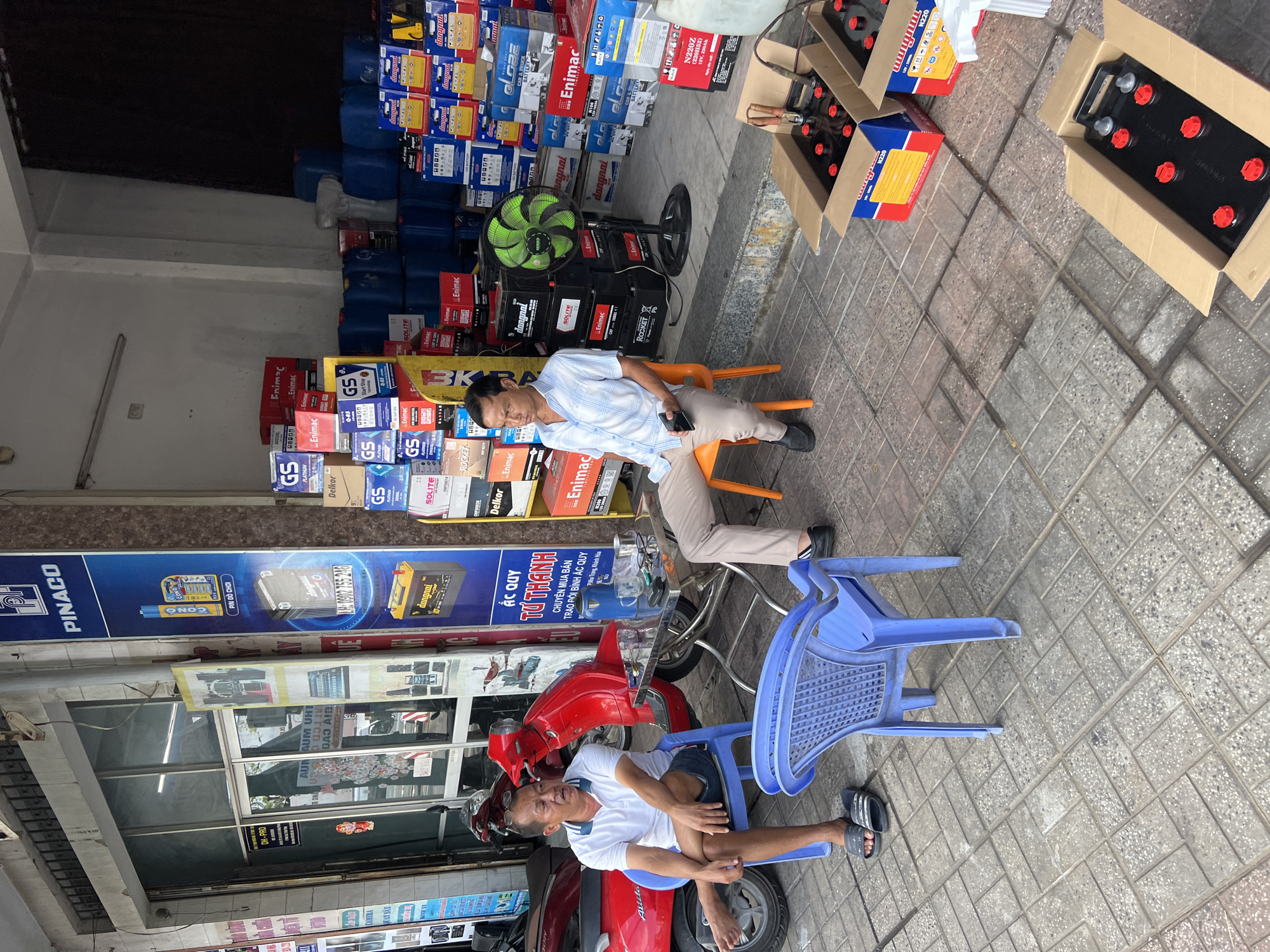 The image depicts a street scene in Nha Trang, Vietnam, likely outside a retail store selling automotive batteries and related products. In the foreground, two men are seated on plastic chairs. One man, wearing a white polo shirt and denim shorts, sits on a blue chair, facing left. The second man, in a light-colored checkered shirt and khaki pants, sits on an orange chair, facing right. A metal table between them holds a blue thermos, a cup, and a pair of glasses. Several red motorcycles are parked behind the man on the blue chair. The background is filled with stacks of car batteries, various containers, and signage indicating the store's offerings, including a prominent "PINACO" sign. A green electric fan is visible among the stacked batteries. The ground is paved with interlocking bricks. The overall impression is of a commercial establishment on a street, with goods displayed both inside and outside the storefront.