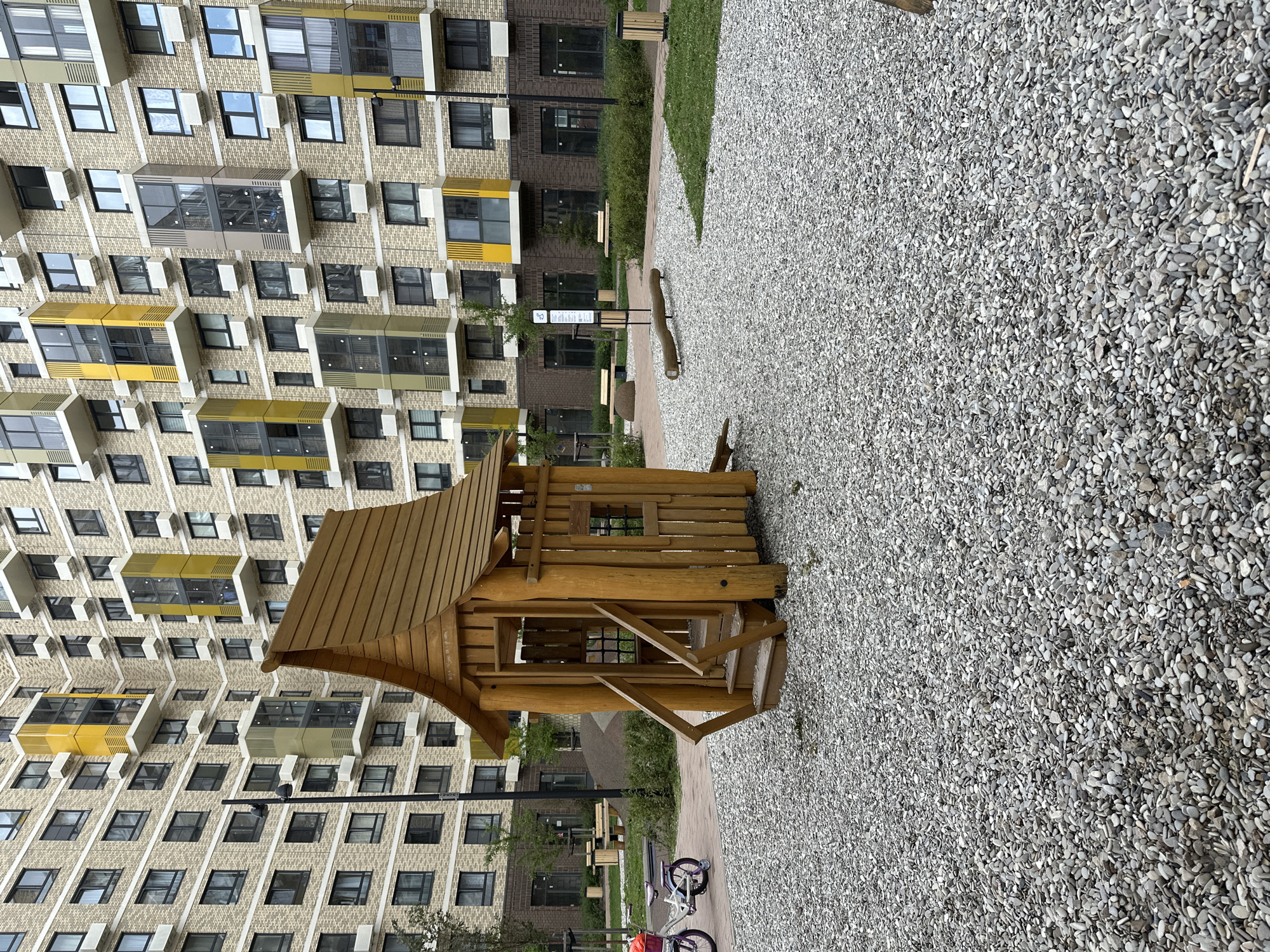 A wooden playhouse with a peaked roof and stairs stands in the foreground on a bed of grey gravel. To the left, a child's bicycle with a pink frame and a basket on the handlebars is parked. In the background, a multi-story residential building with a beige brick facade and yellow and grey accents on balconies is visible. A streetlamp is positioned to the left of the playhouse. Further back, there are benches and green bushes. The scene is set in an outdoor urban environment.