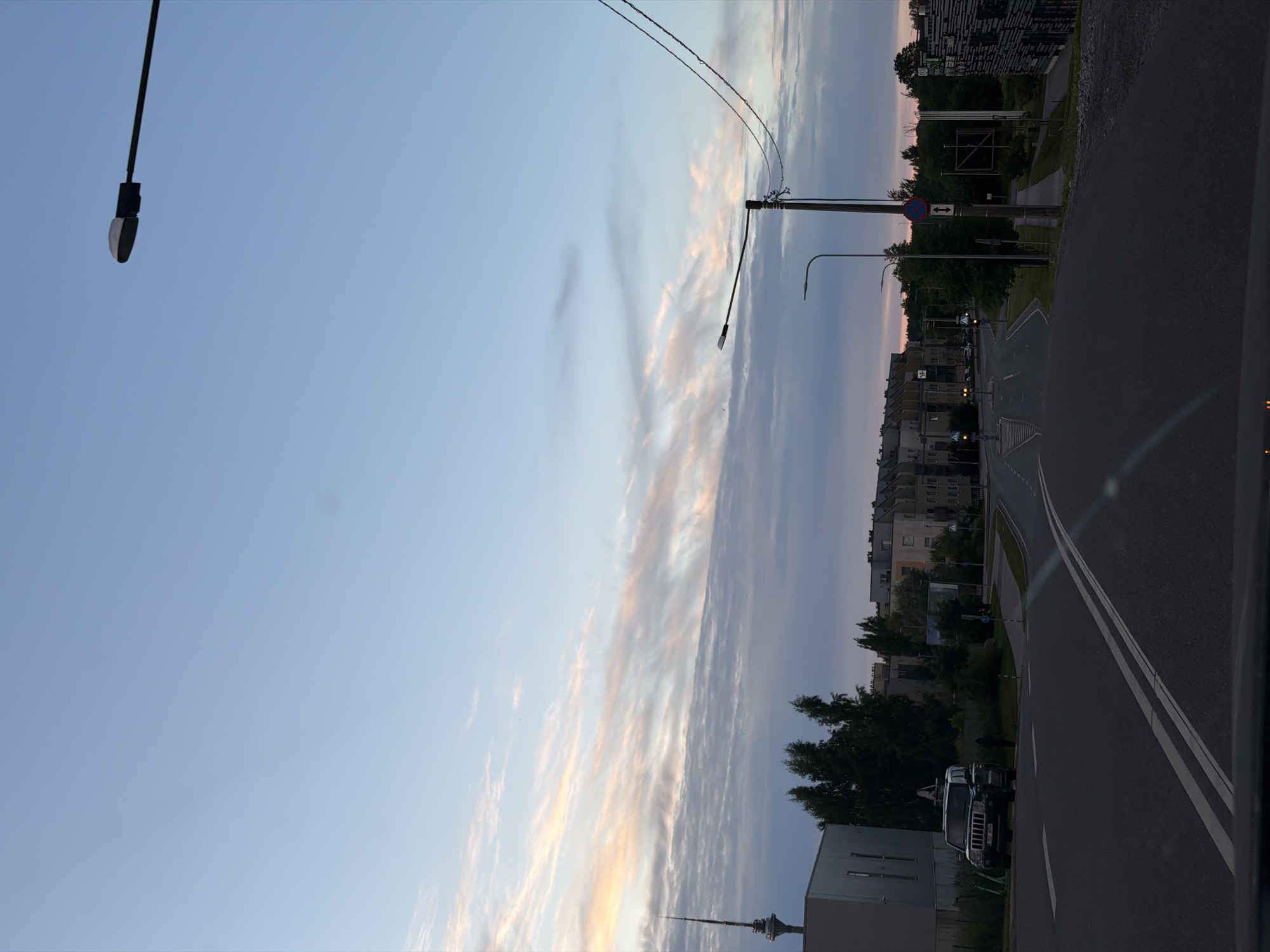 A low-angle shot captures a street scene in Tallinn, Estonia, under a pale blue sky transitioning into twilight. The sky is adorned with wispy, pink-tinged clouds, suggesting the golden hour or early evening. The camera's orientation is vertical, making the scene appear tilted.
In the foreground, a dark asphalt road curves to the right, marked with white lines indicating lanes. To the left of the road, a sidewalk runs alongside a grassy area, bordered by trees and apartment buildings. The buildings are of various heights and architectural styles, with some appearing older and others more modern.
Streetlights are visible, one tall one to the left and another further down the road. Traffic signs are also present, including a circular red and blue sign with arrows, indicating traffic regulations. Several parked cars are visible on the side of the road, including a black SUV.
Further down the road, a slender radio tower or observation tower pierces the skyline, a distinctive landmark. The overall impression is a quiet urban evening scene, with a sense of calm as day transitions to night. No people are clearly identifiable, though figures might be present but indistinct. The weather appears clear, with the clouds being the only notable atmospheric feature. The image conveys a peaceful and mundane slice of urban life.