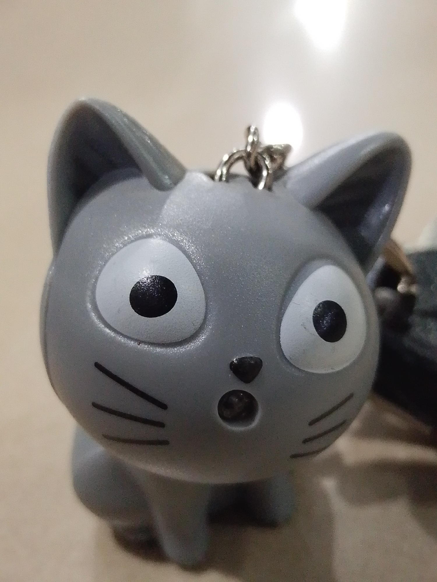 The image is a close-up shot of a gray, cartoon-style cat keychain with large, wide eyes. It features three black whiskers on each side of its face and a small, rounded nose/mouth. A silver keychain ring is attached to the top of the cat's head. The background is a blurred surface, likely a table, with a bright light reflecting on the cat's head. There is no evidence to connect the image to the city of Lhokseumawe, Indonesia.