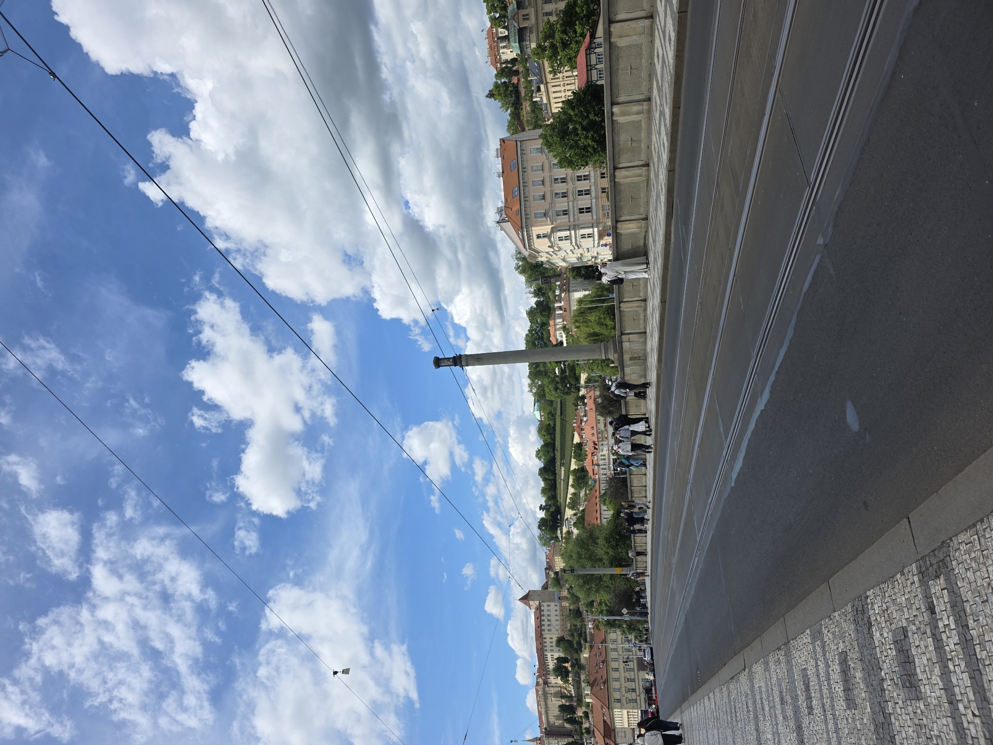 The image shows a view along a city street in an unknown location. The sky is blue with scattered white clouds. Overhead, there are electrical cables. On the right side of the image, there is a pavement with a textured surface. A road runs along the pavement, with tram tracks embedded in the surface. Along the other side of the road, there is a line of buildings, some of which appear to be historical with red roofs. The buildings are interspersed with trees. Several people are visible along the pavement, some are walking. A stone column is visible extending upwards towards the sky.