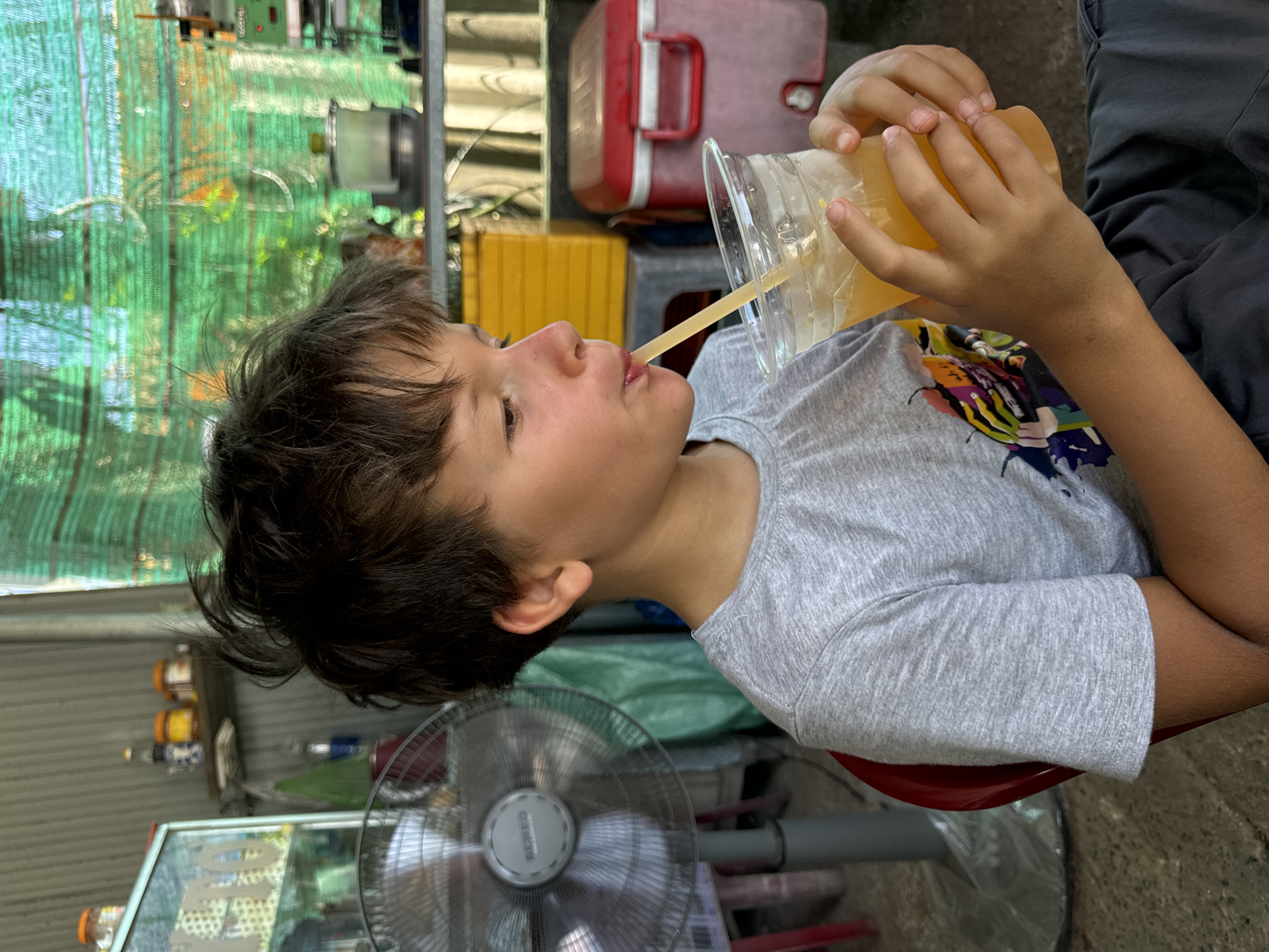 An outdoor scene, possibly in Hồng Sơn, Vietnam, captures a young boy with dark, tousled hair, wearing a grey t-shirt with a colorful graphic print on the left side of the chest. He is seated and holding a clear plastic cup filled with a yellow beverage and ice, drinking through a yellow straw. His hands are positioned around the cup, with his right hand holding it from the side and his left hand supporting the bottom.
In the background, a silver oscillating fan stands to the left of the boy, with its protective grille visible. Behind the fan, a glass display case with items inside is partially obscured. Further back, shelves hold various containers, including yellow and red plastic storage bins. A green mesh material hangs in the background, suggesting an outdoor or semi-covered area. The ground appears to be concrete. The overall impression is a casual, possibly roadside, setting.