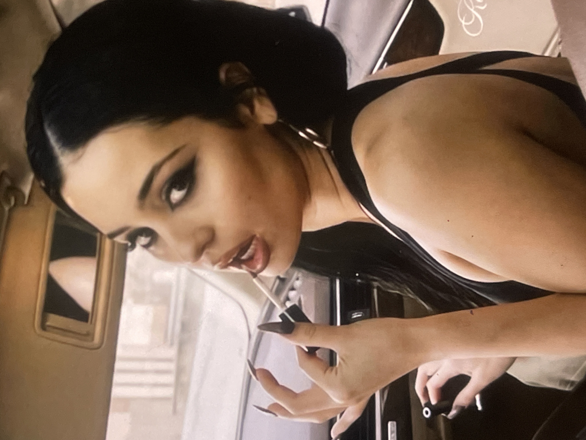 A woman with dark hair pulled back is captured in a close-up, angled shot. She has dramatic eye makeup, featuring sharp winged eyeliner, and is applying lip gloss with a wand. Her lips are slightly parted during the application. She wears a black top with visible straps. Her left hand, with long, dark, pointed nails, holds the lip gloss tube.
The setting appears to be the interior of a vehicle, suggested by visible car elements like a window frame to the left and dashboard or door panels to the right. The background seen through the car window is blurred, showing indistinct shapes that might be buildings or street scenes. In the upper right, a faint, stylized script letter, possibly an "R", is visible on a lighter surface. The woman's expression is focused and direct. This moment of personal grooming is set within the provided location context of Poltava, Ukraine.
