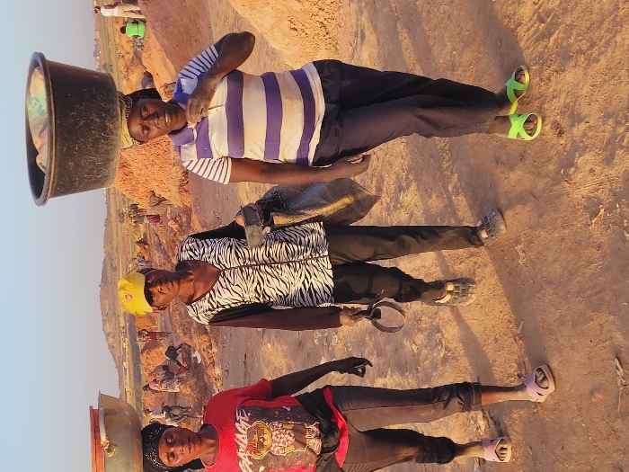 This image captures three adult women walking across a dusty, reddish-brown earthen landscape within the city of Shen, Nigeria. The background reveals numerous other individuals engaged in manual labor amidst piles of excavated earth, suggesting an outdoor work site. The sky is clear.
From left to right:
1. A woman wearing a red t-shirt with a graphic, dark cropped pants, and light pink slide sandals, balances a light-colored, empty basin on her head. She holds a dark object in her right hand.
2. A woman wearing a yellow baseball cap, a black and white zebra-print long-sleeved shirt, dark pants, and dark closed-toe sandals, carries a dark bag on her left arm and holds a dark circular object in her right hand. She looks directly at the camera.
3. A woman wearing a white and light purple/blue striped polo shirt, dark trousers, and bright green slide sandals, balances a dark bucket filled with light-colored material on her head. Her right hand is raised towards her chest, displaying light-colored dust on her fingers.
All three women have dark complexions and neutral expressions. Their primary action is walking while transporting materials or tools, indicative of physical labor in the depicted environment.