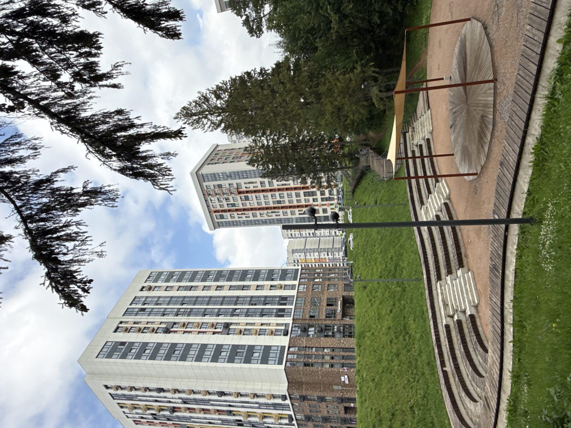An exterior shot, taken from a low angle, depicts a residential complex in Izhevsk, Russia. In the foreground, a semicircular amphitheater made of concrete and wood seating is visible. A circular wooden stage with a beige canopy is situated in the center of the amphitheater. A grassy slope rises behind the amphitheater. In the middle ground, two modern apartment buildings with a mix of white, gray, and brick facades stand tall. One building features colorful vertical accents. Trees and greenery surround the buildings. The sky is cloudy.