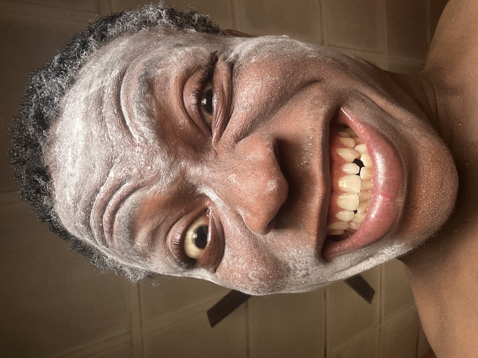 The image depicts a person with dark skin and short, curly black hair. Their face is covered in a white powder. The person is grinning, revealing their teeth. The background appears to be a tiled wall, possibly in a bathroom. The image is taken from a low angle. The city is unknown.