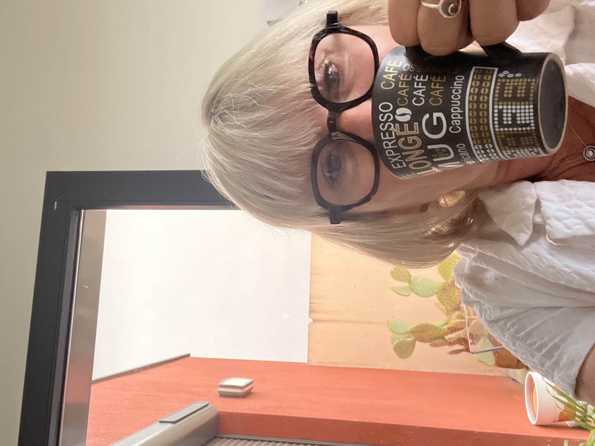 The image depicts a blonde-haired woman with glasses holding a mug that has “EXPRESSO CAFE LONGE MUG Cafe Cappuccino” written on it. The background includes a window frame, an orange wall, a cactus plant, and possibly the sky visible through the window. Based on visual cues, the location appears to be in Chambœuf, France.
