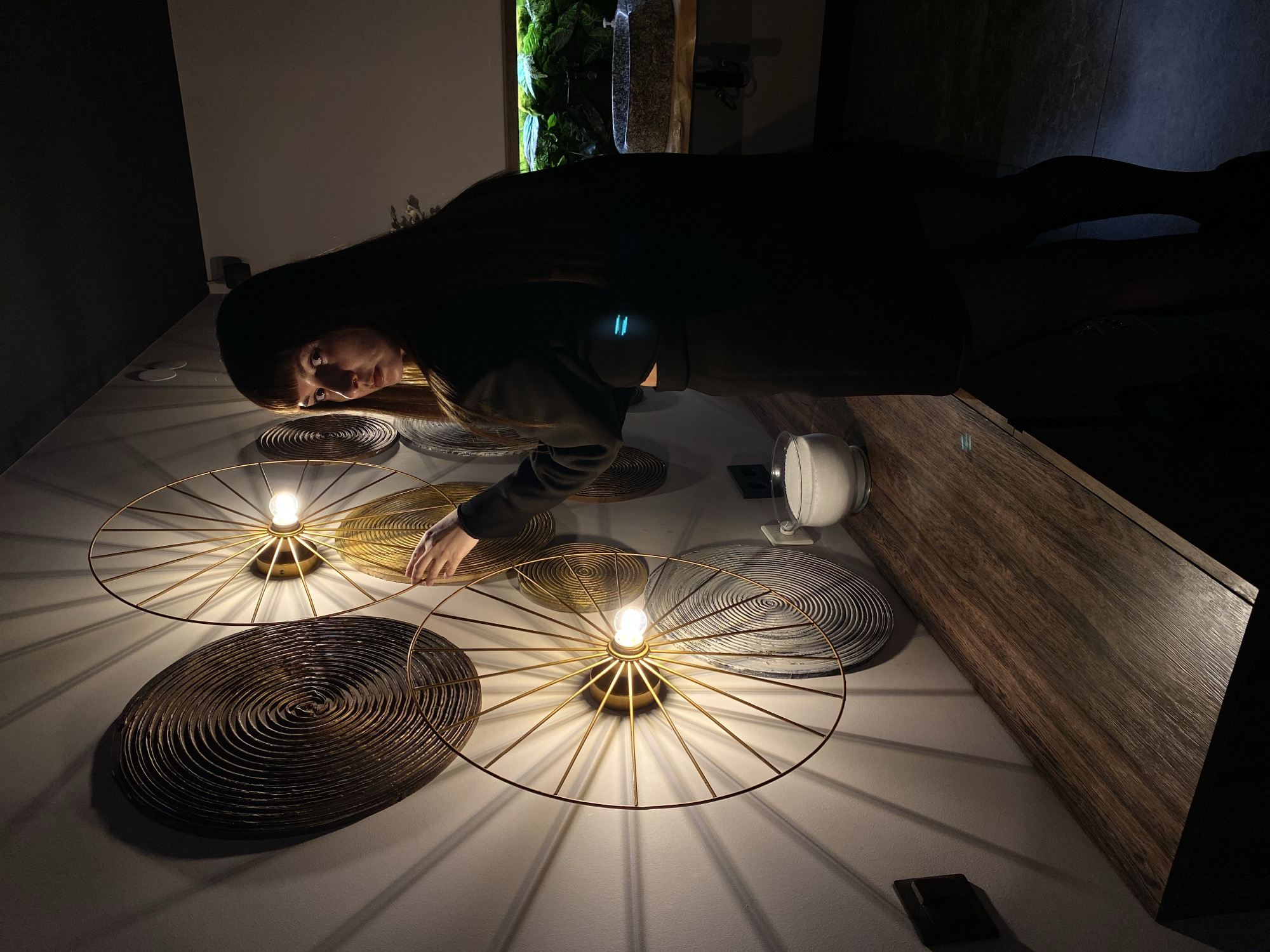 A person with long, dark hair is bent over a series of round, decorative wall lights mounted on a light grey wall. The lights are arranged in a non-uniform pattern. Some lights have brass-colored frames with spoke-like designs emanating from a central bulb. The round decorations within the frames have a textured, spiral pattern. One decoration is brass, another dark bronze, and a third is greyish-white. A white cylindrical object is mounted to the wall nearby. A wooden cabinet is adjacent to the lights.
The location in the city of Votkinsk, Russia cannot be determined.