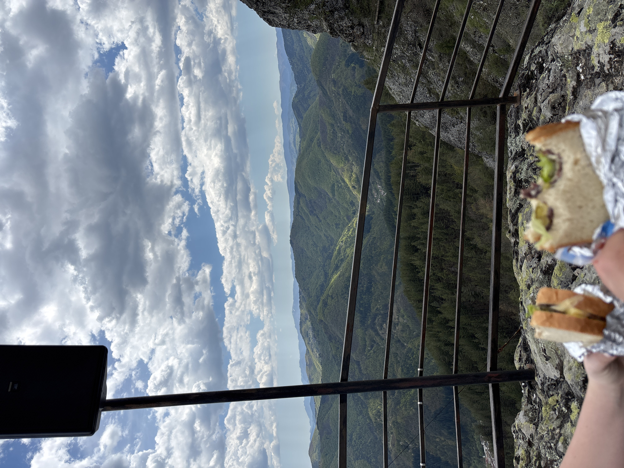 A slightly out-of-focus, rotated, medium shot captures a mountainous vista, likely viewed from a high point near Gradat, Bulgaria. The scene is framed by a dark metal railing running horizontally across the image, and by a rock face with lichen in the upper right corner. A dark sign is visible in the lower left corner. The landscape features rolling hills densely covered in green forest, receding into a hazy blue distance. Above, a sky with mixed white and grey clouds dominates the scene. In the upper right of the shot, someone holds a foil-wrapped sandwich.