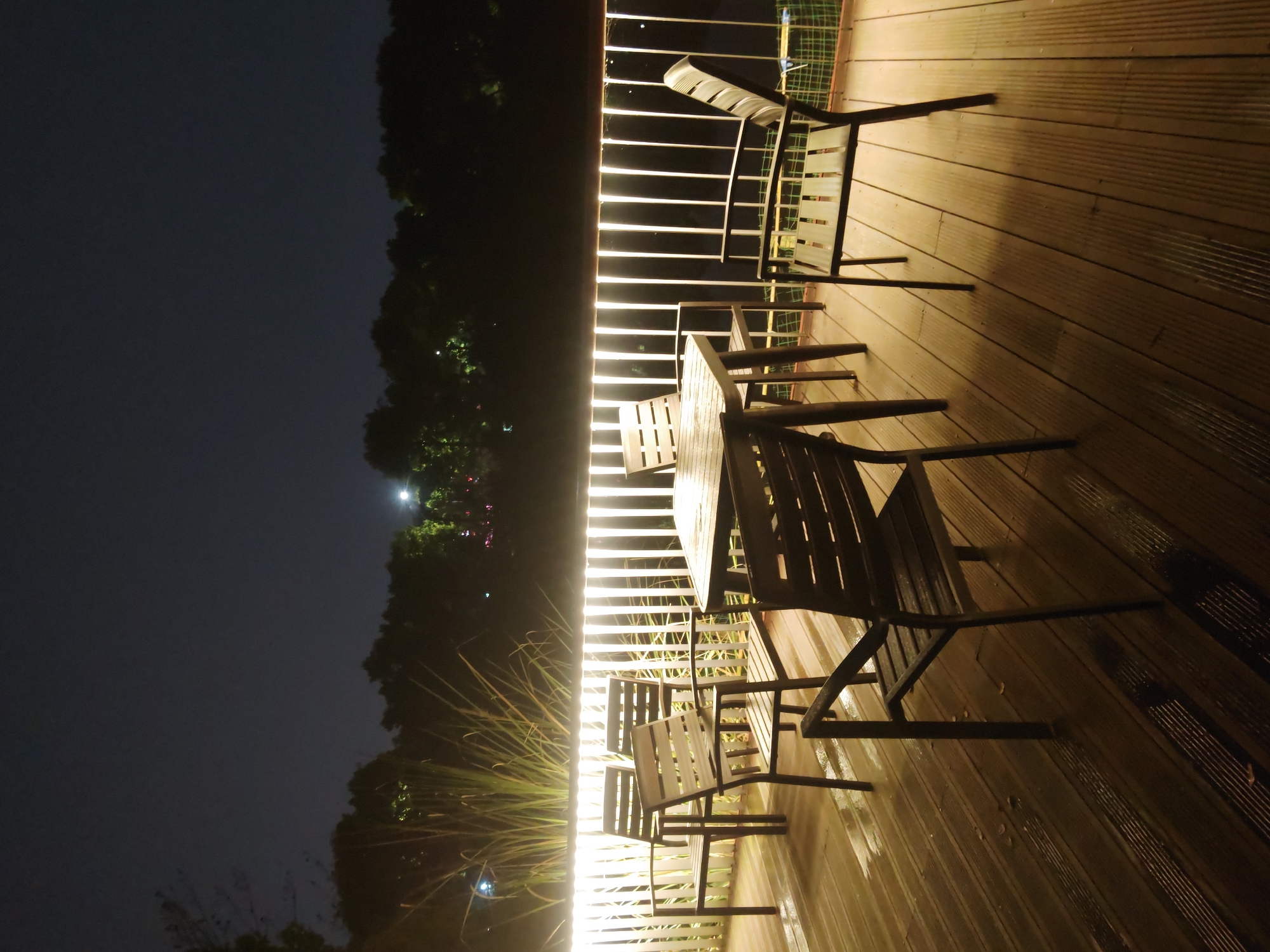The image depicts an outdoor patio at night, likely in Chongqing High-tech Zone, China. The patio has wooden decking and a railing with integrated LED lighting. There are several metal chairs and a small table arranged on the deck. Beyond the railing, there is a dark area with trees, and the night sky. The lighting is bright on the patio, casting long shadows from the furniture. There are no people visible in the scene. The overall environment appears calm and quiet.