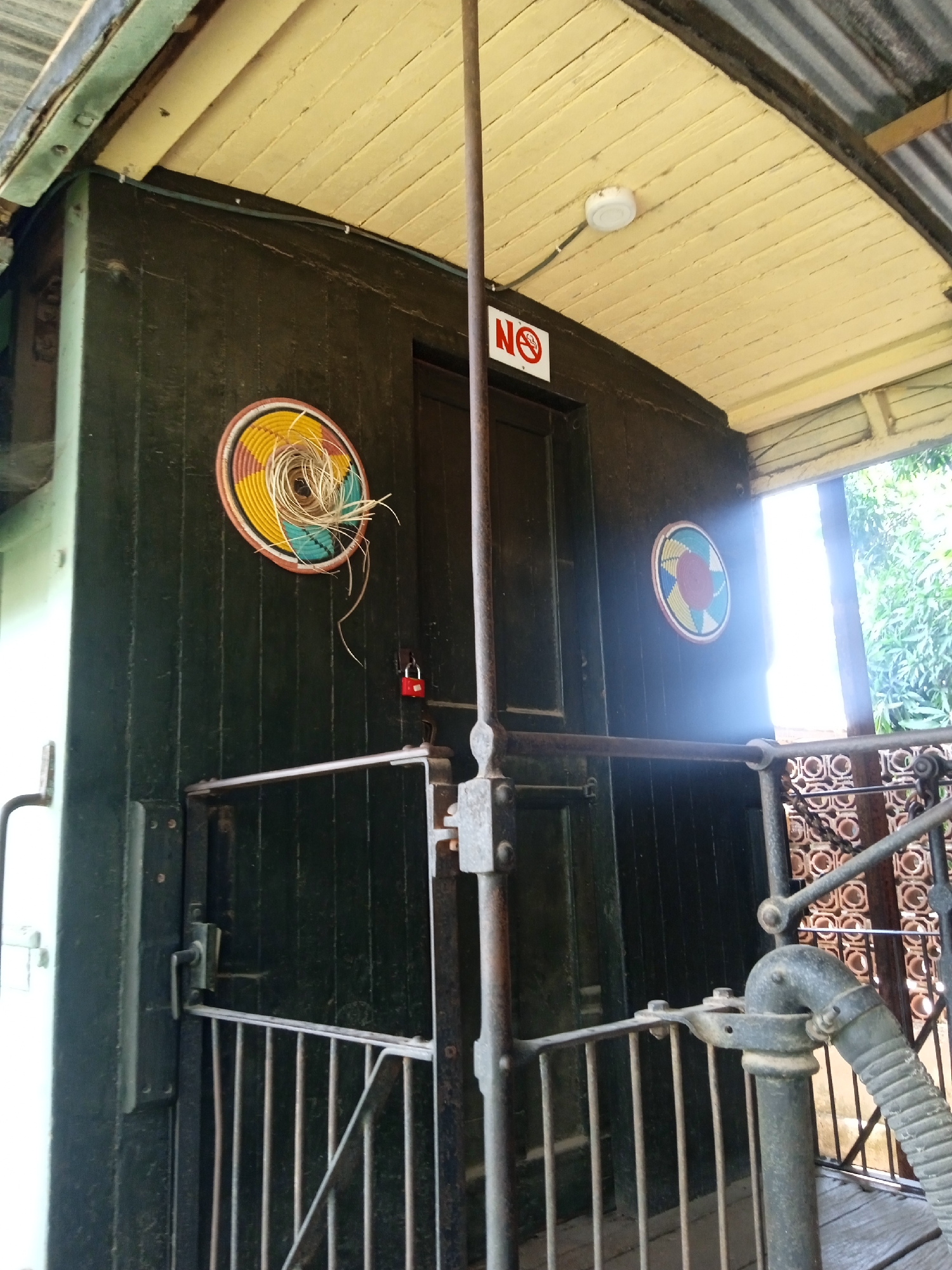 An interior view of a vintage railway carriage is seen. The carriage is paneled with dark wood and has a cream-colored ceiling. On the left side of the carriage, a decorative circular basket with multiple strands of a plant sticking out of it is visible, secured with a red padlock, likely signifying that it is locked. A smaller, similar basket hangs on the right side. A metal gate with vertical bars is in the foreground, partially obscuring the view into the carriage. An "NO" sign is affixed above a door. Light filters in through a window on the right. The environment outside the window includes a leafy tree and a patterned wall. This scene is most likely a preserved historical exhibit in Jos, Nigeria.