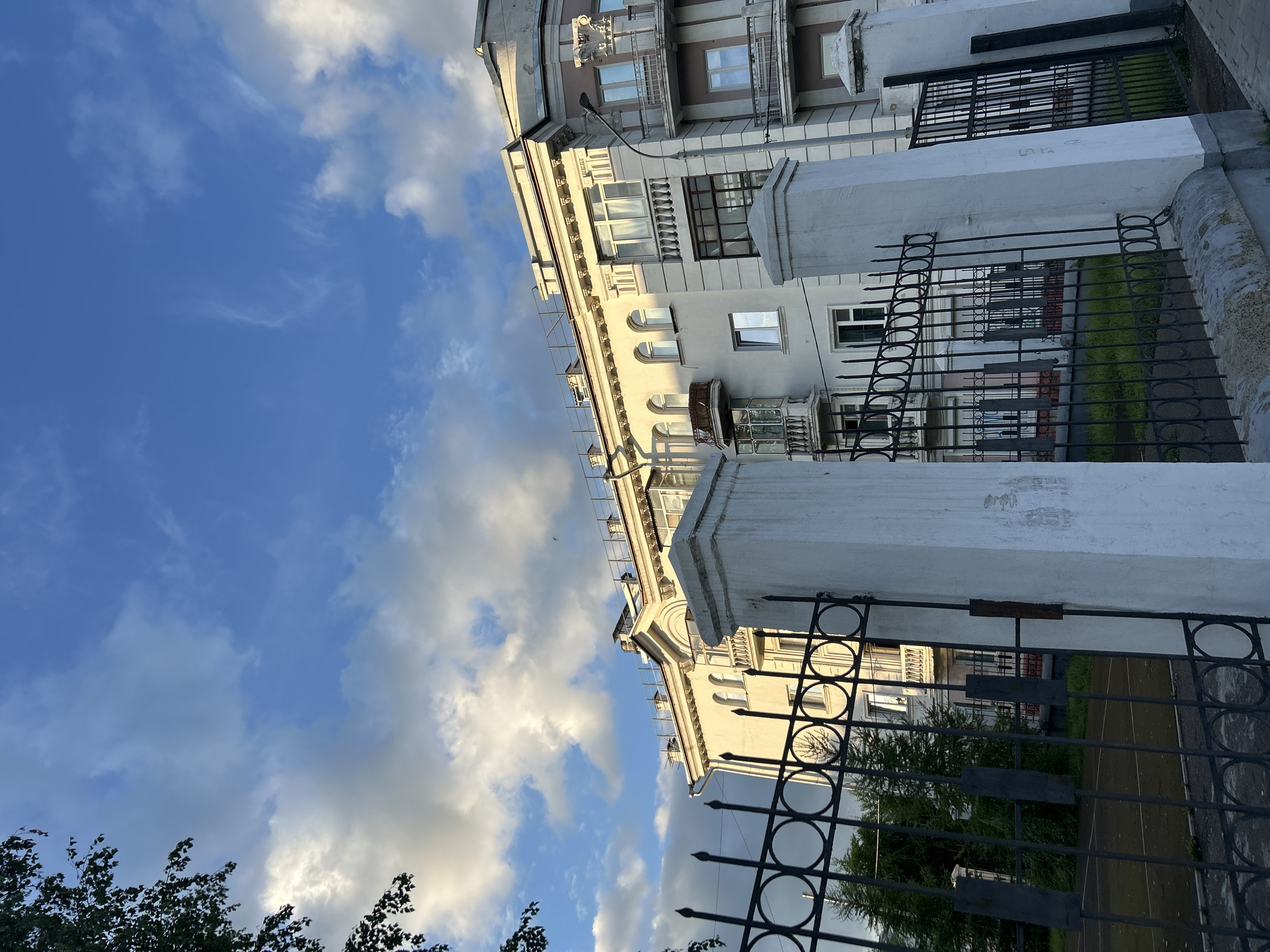 A low-angle exterior shot captures a portion of an ornate, light-colored building against a dynamic sky filled with white and gray clouds. The building features multiple stories with arched windows, balconies with decorative railings, and classical architectural elements like columns and cornices. In the foreground, a black wrought-iron fence with a circular pattern and pointed finials runs horizontally across the frame, partially obscuring a glimpse of green grass and trees behind it. Two substantial white concrete pillars with a textured surface stand on either side of the fence. The lighting suggests it is late afternoon or early evening, with sunlight casting shadows and highlighting the building's details.