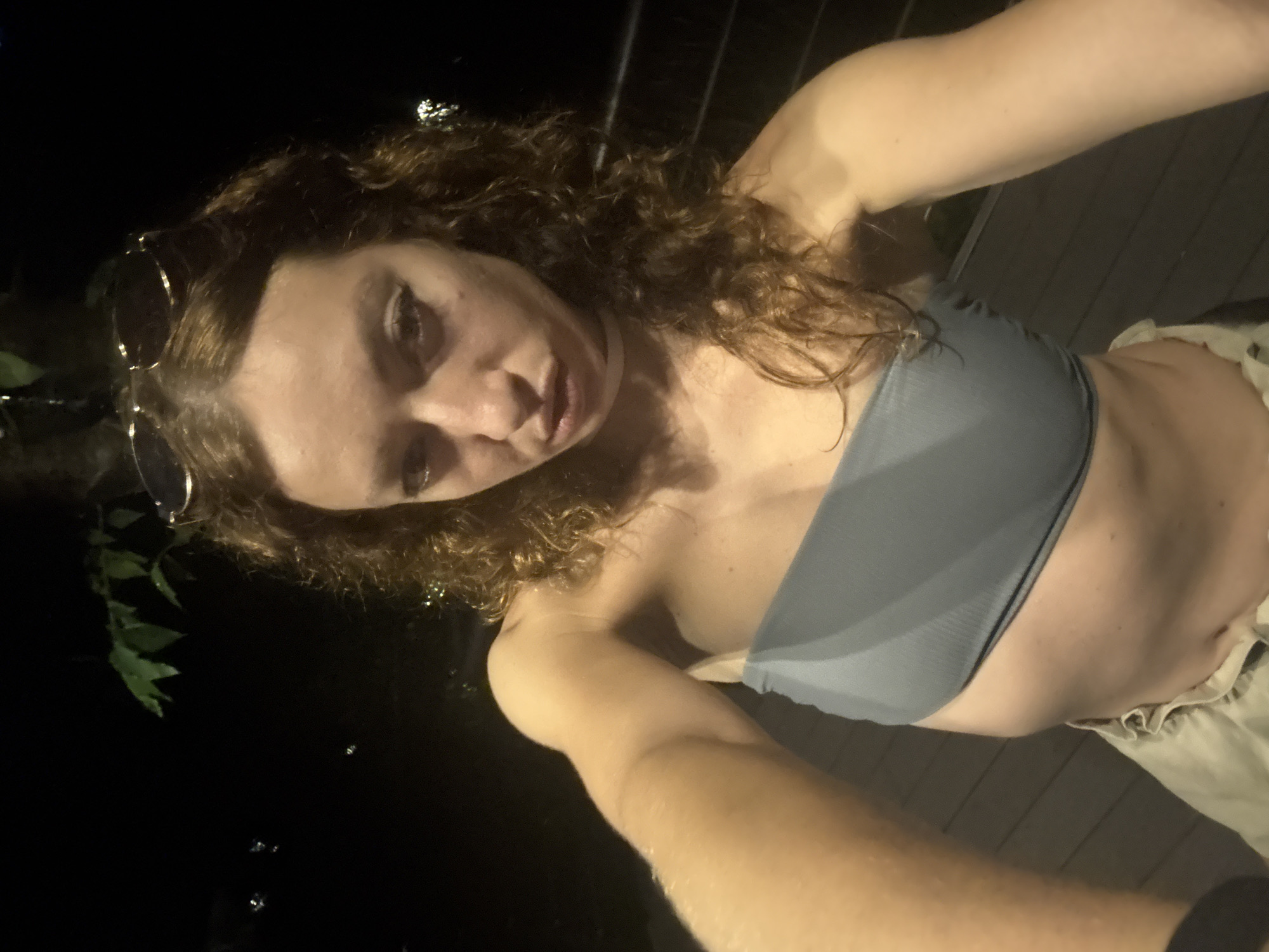 A young woman is captured in a nighttime selfie, illuminated by a direct light source against a dark background. She has curly, reddish-brown hair, with sunglasses resting on her head and a large hoop earring visible on her left ear. She wears a light grey strapless bandeau top and light-colored shorts or pants, visible at her waist. Her arms are extended, holding the camera from an overhead perspective, and she looks directly at the viewer with a neutral to slightly confident expression.
In the background, dark, blurred green foliage is visible on the left side. A horizontal slatted structure, possibly a railing or decking, runs along the right side and behind her, suggesting an outdoor setting like a balcony or patio. Faint, indistinct lights twinkle in the distant darkness, hinting at a view over a populated area or body of water. The sharp contrast between the well-lit subject and the deep shadows of the background indicates the use of a flash or other direct light source. No text is visible in the image.