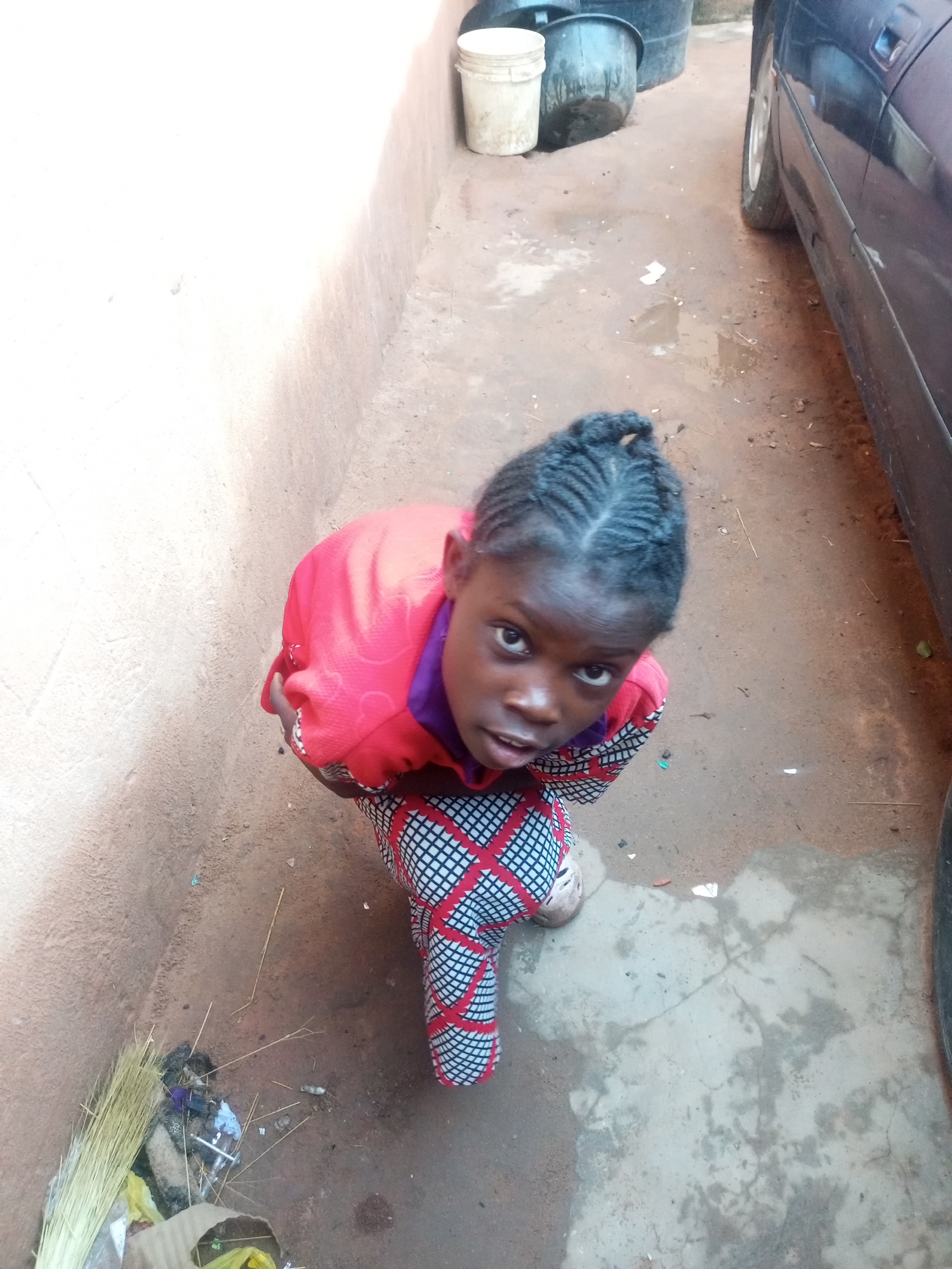 A young Black female, appearing to be a child, is kneeling on a dirt ground next to a light-colored wall. She is looking up at the camera. She has her hair braided in a style that is pulled back from her face. She is wearing a red long-sleeved shirt with a purple collar and a black and white checkered skirt. Her feet are partially visible, with one foot wearing a beige shoe and the other wearing a dark shoe.
In the background, a dark blue vehicle is parked to the right of the frame. To the left of the vehicle are several large black plastic containers and a white plastic bucket. The ground appears to be a mixture of dirt and some concrete. There are some scattered pieces of trash on the ground, including a broom leaning against the wall and some yellow and white plastic bags. The scene suggests an outdoor environment, possibly a residential area or a yard, in Tempure, Nigeria.