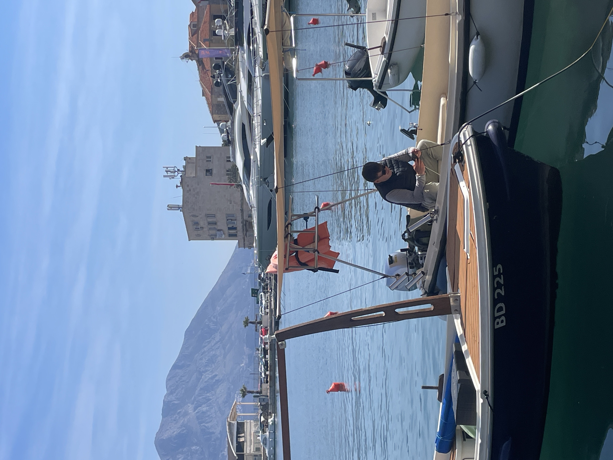 The scene depicts a marina in Budva, Montenegro, under a clear blue sky with a large, rugged mountain range in the background on the left. In the foreground, a dark blue boat with "BD 225" stenciled on its hull, featuring a wooden deck and a brown wooden mast structure, is moored. To its right, a male individual is seated facing downward in a light-colored boat. He is wearing sunglasses, a dark vest over a light long-sleeved shirt, and light-colored trousers, and is actively engaging with a small electronic device held in his hands. This boat is equipped with a Yamaha outboard motor at its stern and an orange life vest hanging from an overhead canopy frame. Another white boat, partially visible to the right, also has an outboard motor and a white fender attached to its side. Orange buoys are visible floating in the blue water further in the midground. The background reveals several buildings along the shore, including a multi-story, light-colored building with multiple communication antennas on its roof, and traditional-style buildings with terracotta roofs. Numerous large, luxurious yachts are moored in the distance, alongside a parking area with parked cars and palm trees.