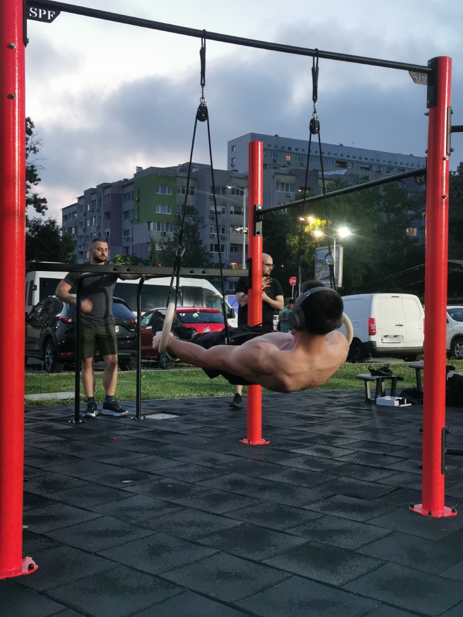 An outdoor calisthenics park in Burgas, Bulgaria, is depicted at dusk or early evening under an overcast sky. In the foreground, a shirtless, muscular male wearing dark shorts and over-ear headphones is suspended horizontally from white gymnastic rings, performing what appears to be a front lever. He is facing away from the camera, demonstrating significant upper body strength. The rings are attached by black straps and pulleys to a black horizontal bar supported by thick red vertical posts, part of a fitness structure featuring "SPF" branding.
To the left of the central figure, a man with a short beard, wearing a dark grey t-shirt, dark shorts, and athletic shoes, stands leaning on a lower horizontal bar of the structure, observing the exercise. To the right, a bald man with glasses and a beard, wearing a dark t-shirt with visible arm tattoos, also stands observing the man on the rings.
The ground of the park is covered with dark, rectangular rubber safety tiles, some appearing wet. In the background, several multi-story residential buildings with numerous windows and air conditioning units are visible, including one with a distinctive green section. Vehicles parked behind the exercise area include a dark SUV, a white panel van, and another white panel van displaying a Bulgarian license plate "A 59 PB," which corresponds to the Burgas region. Streetlights in the background are illuminated, casting bright glows. A red "STOP" sign is partially visible behind the bald observer. A small black stool or bench with a white box rests near the right red support post.