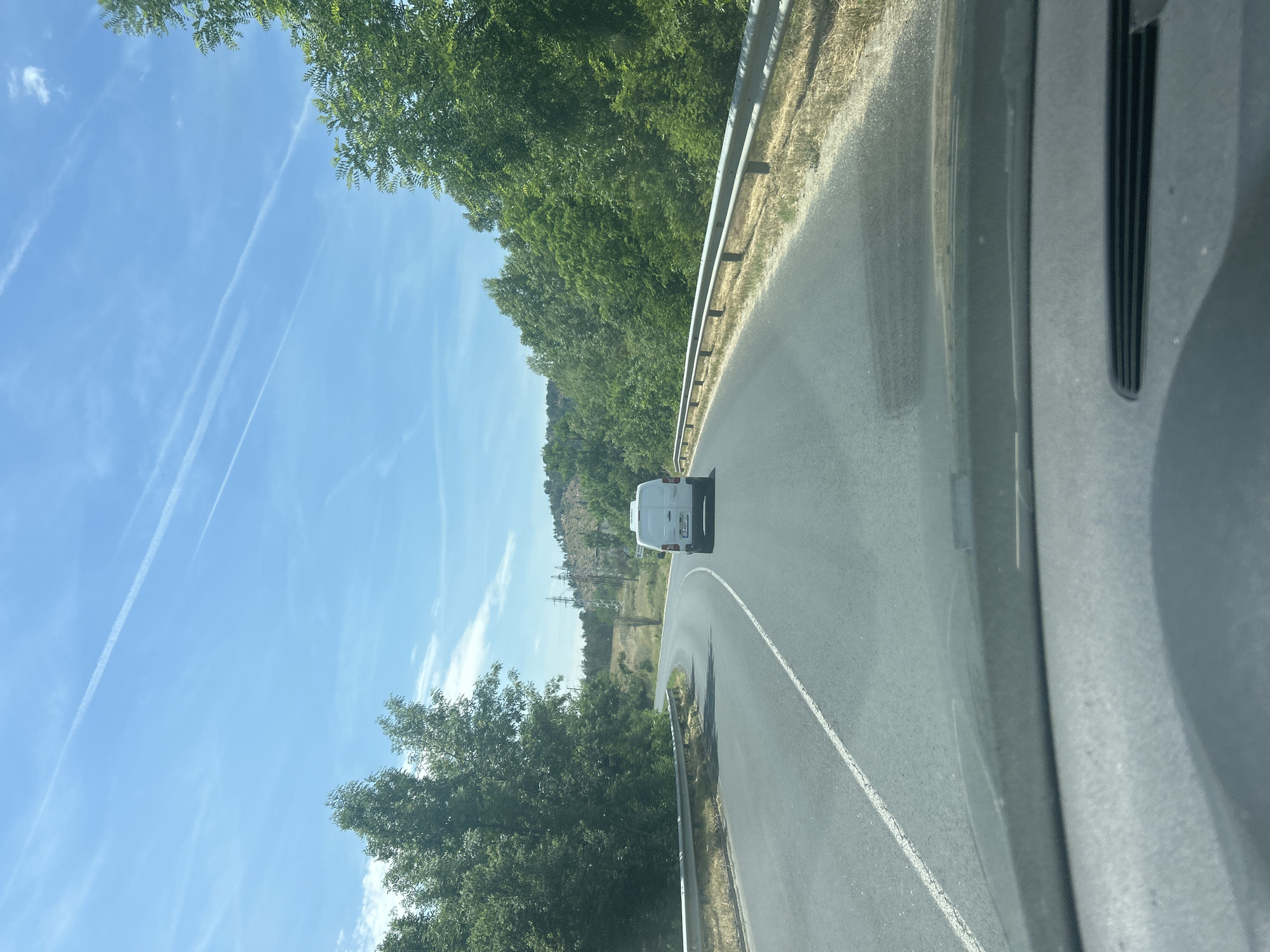 The image captures a bright daytime scene from the perspective of a vehicle, looking forward at a winding asphalt road. A white van is visible ahead, driving on the right side of the road as it curves to the left. The sky is clear and blue with several prominent white contrails stretching across it.
The road features a solid white line on the right shoulder and a broken white line marking the center as it follows the curve. A silver metal guardrail runs alongside the right edge of the road. Lush green trees line both sides of the road, forming dense foliage. In the mid-ground on the left, beyond the trees, there are power lines or utility pylons visible against a backdrop of a drier, more open hillside. Further in the distance, rolling hills or mountains are discernible under the bright sky.
The immediate foreground on the right side of the image shows parts of the interior of the car from which the photo was taken, including what appears to be a portion of the dashboard or windshield frame. The time of day is sunny, likely midday, with strong natural light. The overall setting suggests a journey through a rural or semi-rural landscape. No discernible text is clearly legible, including on the van's license plate.