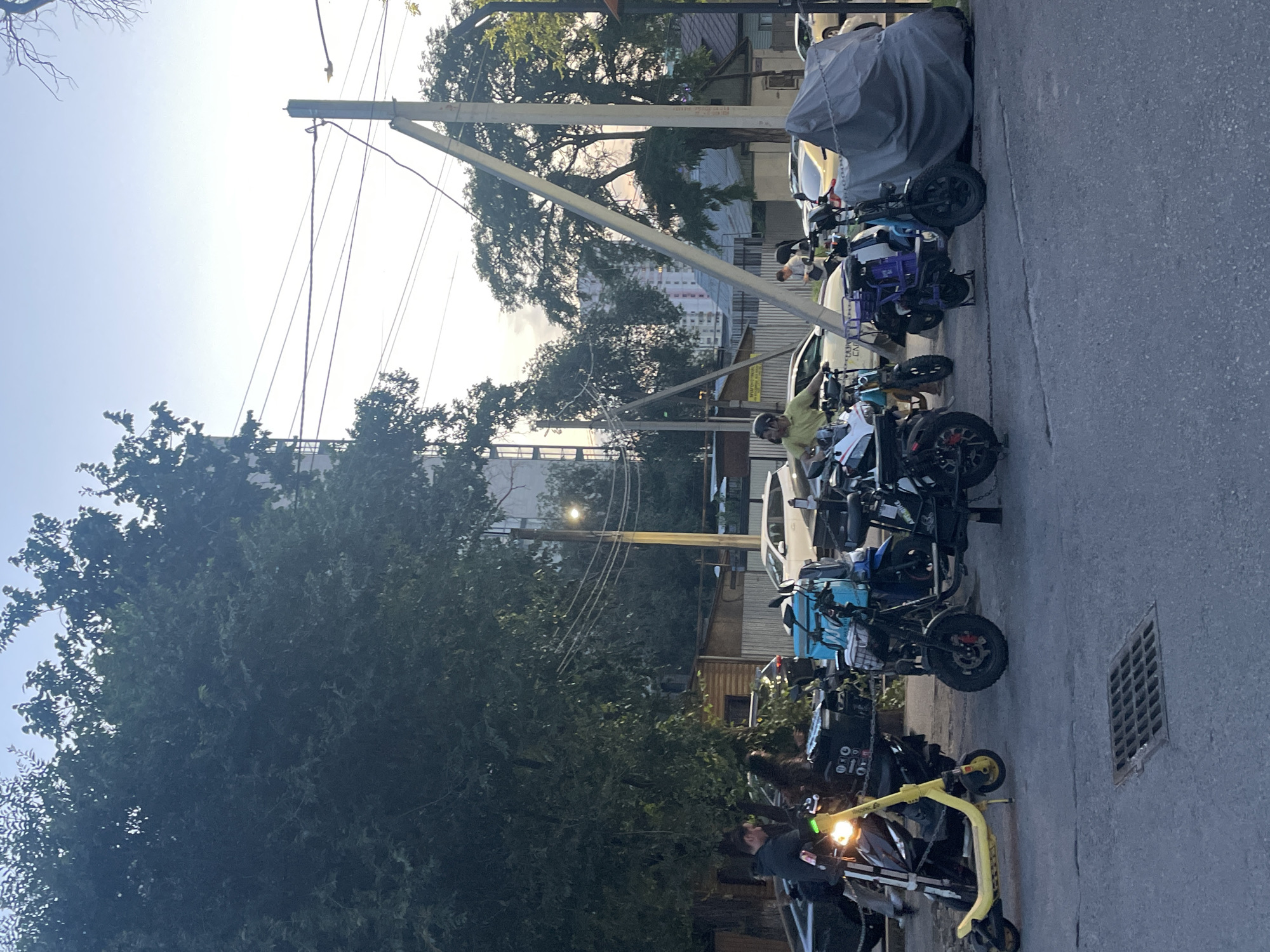 The image depicts an outdoor scene in Almaty, Kazakhstan, characterized by a group of parked electric scooters and motorcycles. Several individuals are present, some near the vehicles. The environment includes a paved road with a storm drain grate in the foreground, surrounded by trees, a building, and utility poles with overhead wires. One scooter in the foreground is yellow, and others are black, blue, and purple. One black motorcycle is covered by a grey tarp. The sky is clear and light blue, suggesting it is either early morning or late afternoon.
