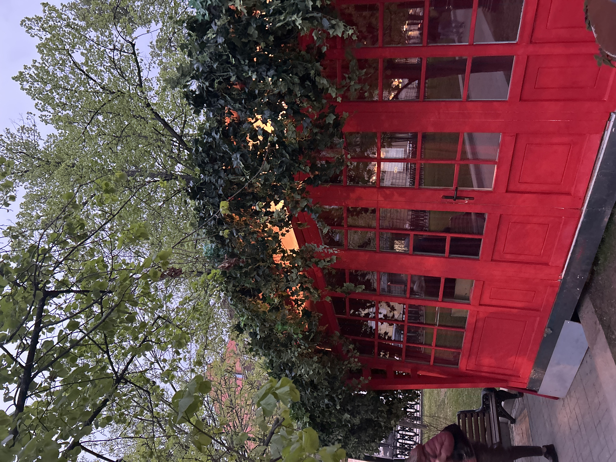A vibrant red structure, reminiscent of a vintage telephone booth, is partially covered in lush green ivy. The structure has multiple window panes reflecting the surrounding environment. A tree with light green foliage is nearby. Below the red structure, a park bench is situated on a paved surface, and a person, partially visible, appears to be walking. The location is presumed to be a city based on the urban setting and architecture in the window reflections.