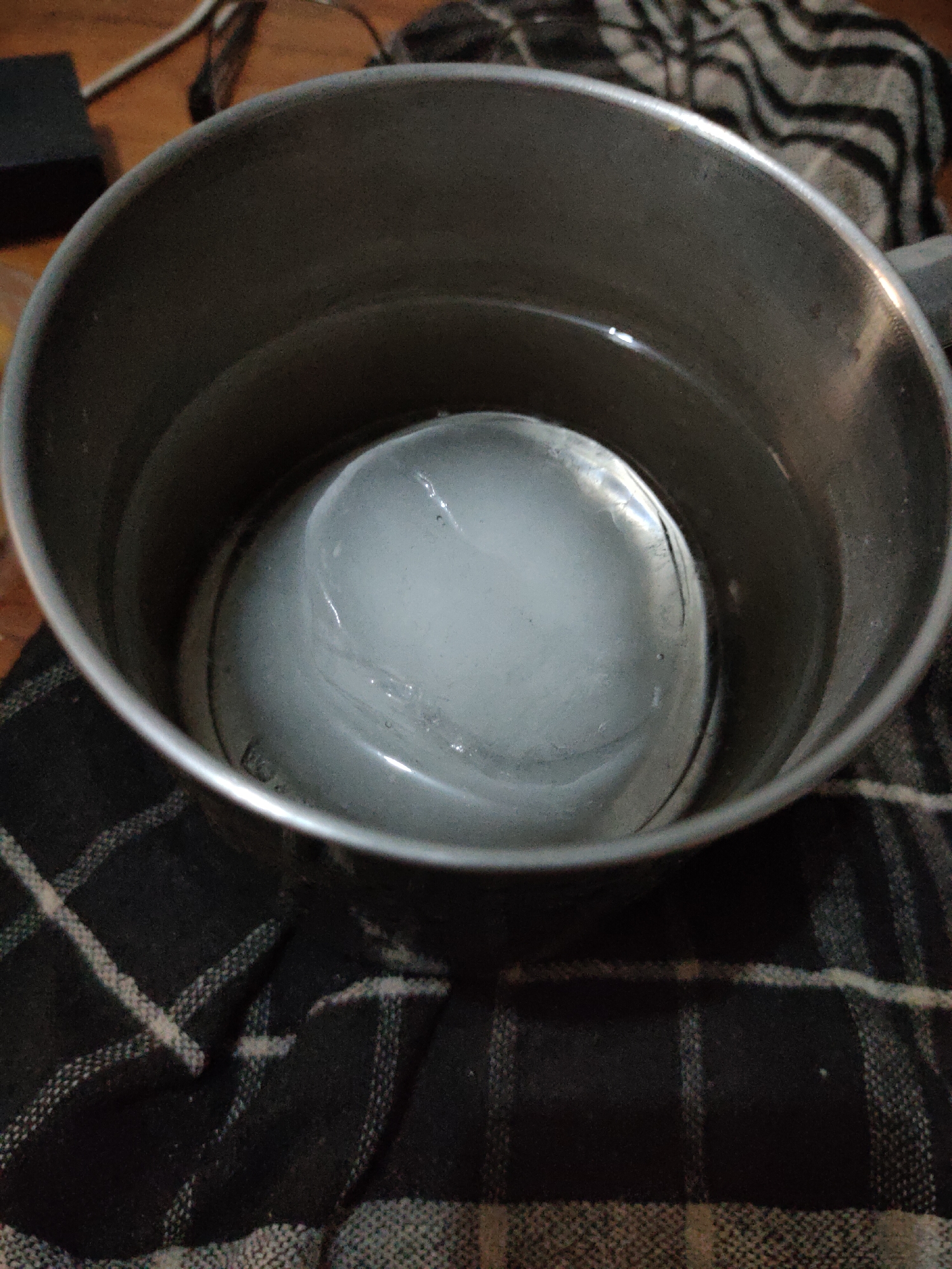 A close-up, high-angle shot shows a large, spherical ice ball suspended in water within a metal pot. The pot is placed on a dark fabric with a checkered pattern. The ice ball is translucent, revealing some internal structures and slight imperfections. The water is clear, and the interior of the pot appears to be metallic and slightly worn. The background is blurred, showing some wooden surfaces and electrical cords, suggesting an indoor environment. The image lacks any specific geographic markers that would place it in South Jakarta, Indonesia. The scene appears to be a still life composition focusing on the ice and water.