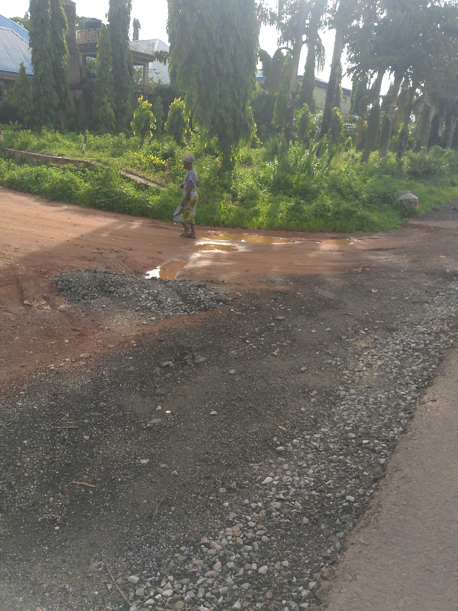 The image depicts a scene on a dirt road in Keffi, Nigeria. A woman wearing a patterned wrap dress and headscarf walks on the muddy road, stepping over puddles of water. In the foreground, the road is covered with gravel and dirt, indicating some form of road work or repair. Behind the woman, lush green vegetation lines the side of the road, with tall trees and some partially visible buildings in the background. The overall impression is of a rural or semi-urban environment within Keffi, with evidence of recent rainfall contributing to the wet road conditions.