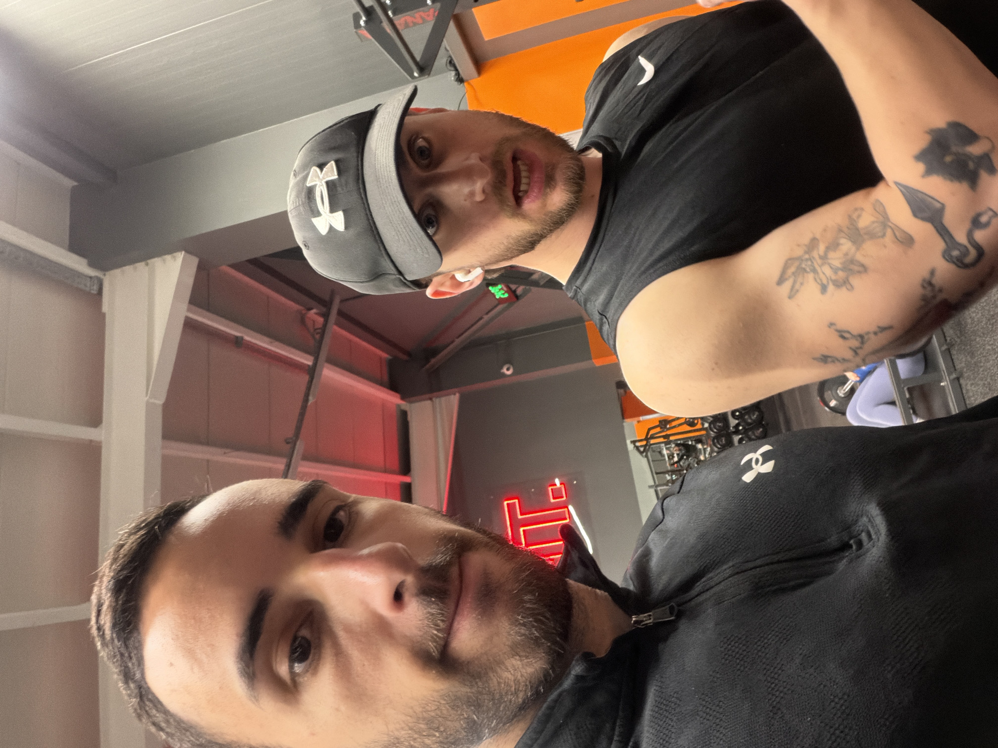 Two men are in a gym. The man closer to the camera has dark hair and a beard, wearing a black Under Armour zip-up jacket. The other man is wearing a black Under Armour t-shirt and a grey Under Armour baseball cap. He has tattoos on his upper arm. Gym equipment and weights are visible in the background. A red neon sign is illuminated on the wall. The location is likely in Plovdiv, Bulgaria, given the prompt.