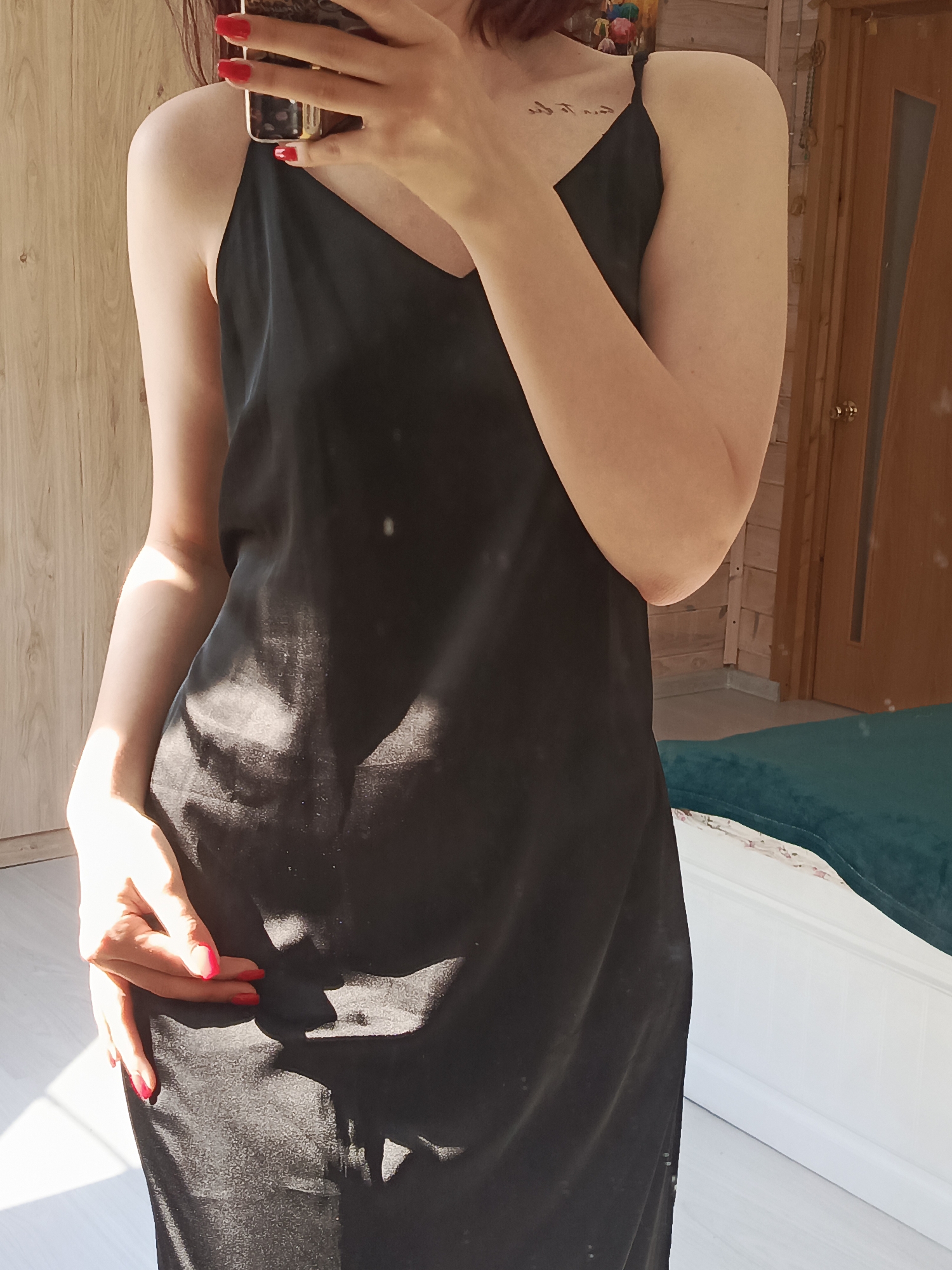 A person is standing indoors, wearing a black dress with spaghetti straps, and taking a selfie with a mobile phone. The person has red nail polish. The room has light-colored wooden walls, a wooden door, and a piece of furniture with a green cushion. The location is unknown and cannot be determined from the image.