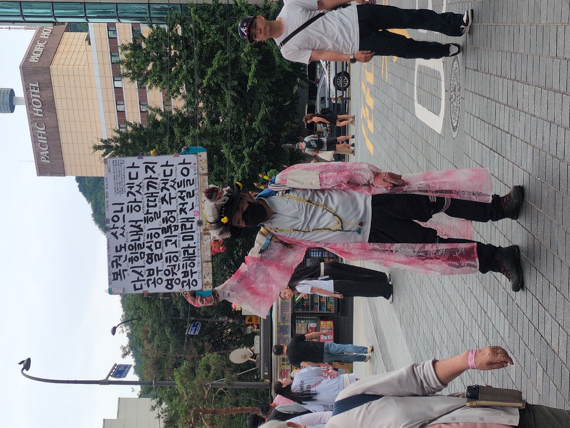 This image captures a daytime urban street scene in Seoul, South Korea, specifically near the Pacific Hotel (now L'Escape Hotel) at the base of Namsan Mountain, with the N Seoul Tower visible in the background.
In the center of the frame, a person stands facing slightly left, holding a large white rectangular sign overhead. This individual wears a black face mask, a white long-sleeved shirt, black pants, and dark footwear. Over their shirt, a translucent, light pink/white long robe with red striped accents is draped. A multi-colored, elaborate headpiece adorned with small yellow duck figures and fabric scraps is worn. A necklace of beads hangs around their neck. The sign they hold displays Korean text in black lettering, which translates to: "I even bought a lottery ticket / I will gather strength again / Until I study diligently / You tormented me forever / Study. Future legends." Smaller handwritten notes also appear around the main text.
To the right of the central figure, a man in a white t-shirt, black pants, and a dark baseball cap with a white "A" logo walks towards the viewer's right. A black strap crosses his chest. On the far left, a portion of a person wearing a light grey long-sleeved top with a pink wristband and a tan shoulder bag is visible. Several other pedestrians are scattered in the mid-ground and background, walking on the paved street and sidewalks. One woman in a white t-shirt and dark pants stands near a small commercial stall in the mid-left background.
The background features the prominent "PACIFIC HOTEL" building, a multi-story structure with light-colored walls and dark brown upper sections, with the N Seoul Tower visible on a distant hill behind it. Another modern building with green reflective glass panels is partially seen on the far right. Streetlights, a pole with a satellite dish, and blue Korean street signs are visible. The ground is paved with rectangular grey and brown tiles, featuring yellow painted lines and a white '0' or 'D' shaped marking, along with a manhole cover. Lush green trees and vegetation line the street and cover the hills in the distance. The sky is overcast, providing diffused lighting to the scene.
