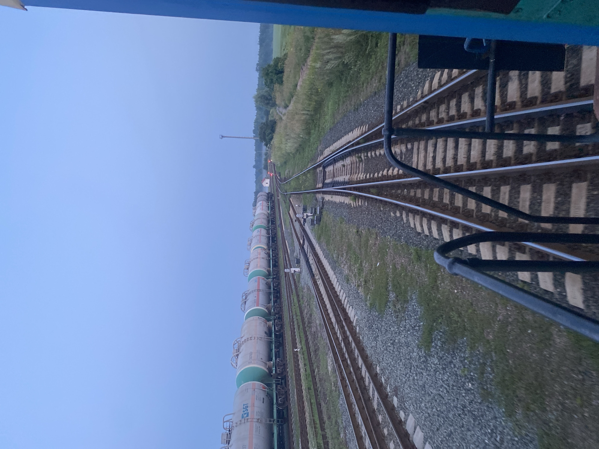 The image shows a train consisting of multiple tanker cars stopped on a railway line at Kauchuk Stantsiya, Russia. The sky is a pale blue, suggesting it is either early morning or late evening. The train cars are cylindrical and appear to be transporting liquids. They are parked on a set of parallel railway tracks, with ballast and grass visible around the tracks. In the background, there is a field, trees, a small building, and a tall tower, possibly a communication tower or a signal tower. The perspective is from a point on another train or structure, looking forward along the tracks. There are no people visible in the image. The overall scene depicts a quiet, industrial setting at a railway station.