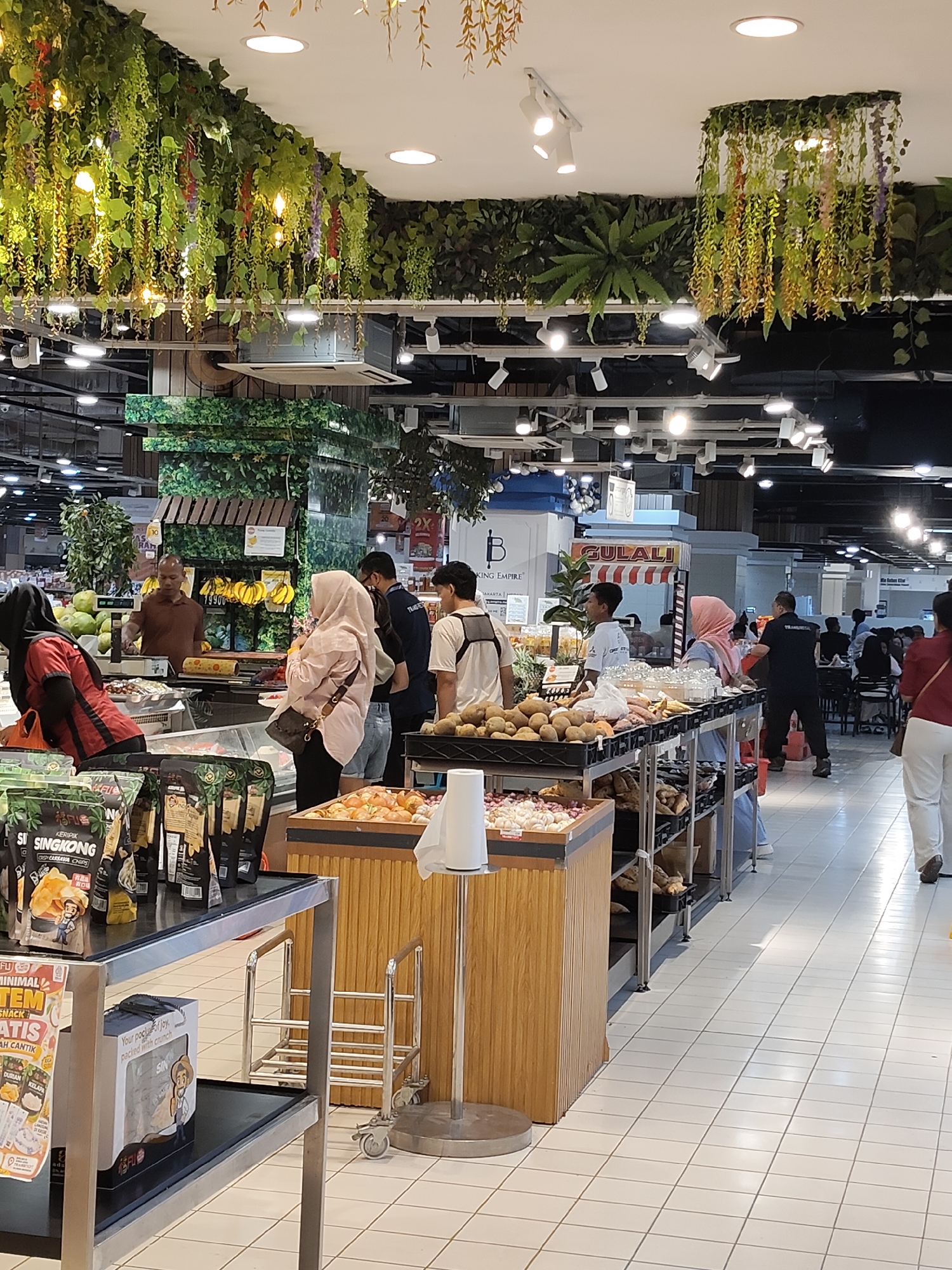 This is a street-level, eye-level shot of a busy supermarket produce section in Medan, Indonesia. In the foreground, a metallic display shelf holds packaged snacks, with a white paper roll dispenser to the right. A wooden display cart filled with onions and potatoes is positioned next to it. The background reveals a diverse range of shoppers and staff interacting with produce displays, including fruits and vegetables. The supermarket is decorated with artificial greenery hanging from the ceiling and walls, creating a lush, natural ambiance. A food stall with a banner reading "GULALI" is visible further back, indicating a food service area. The floor is tiled with white squares.