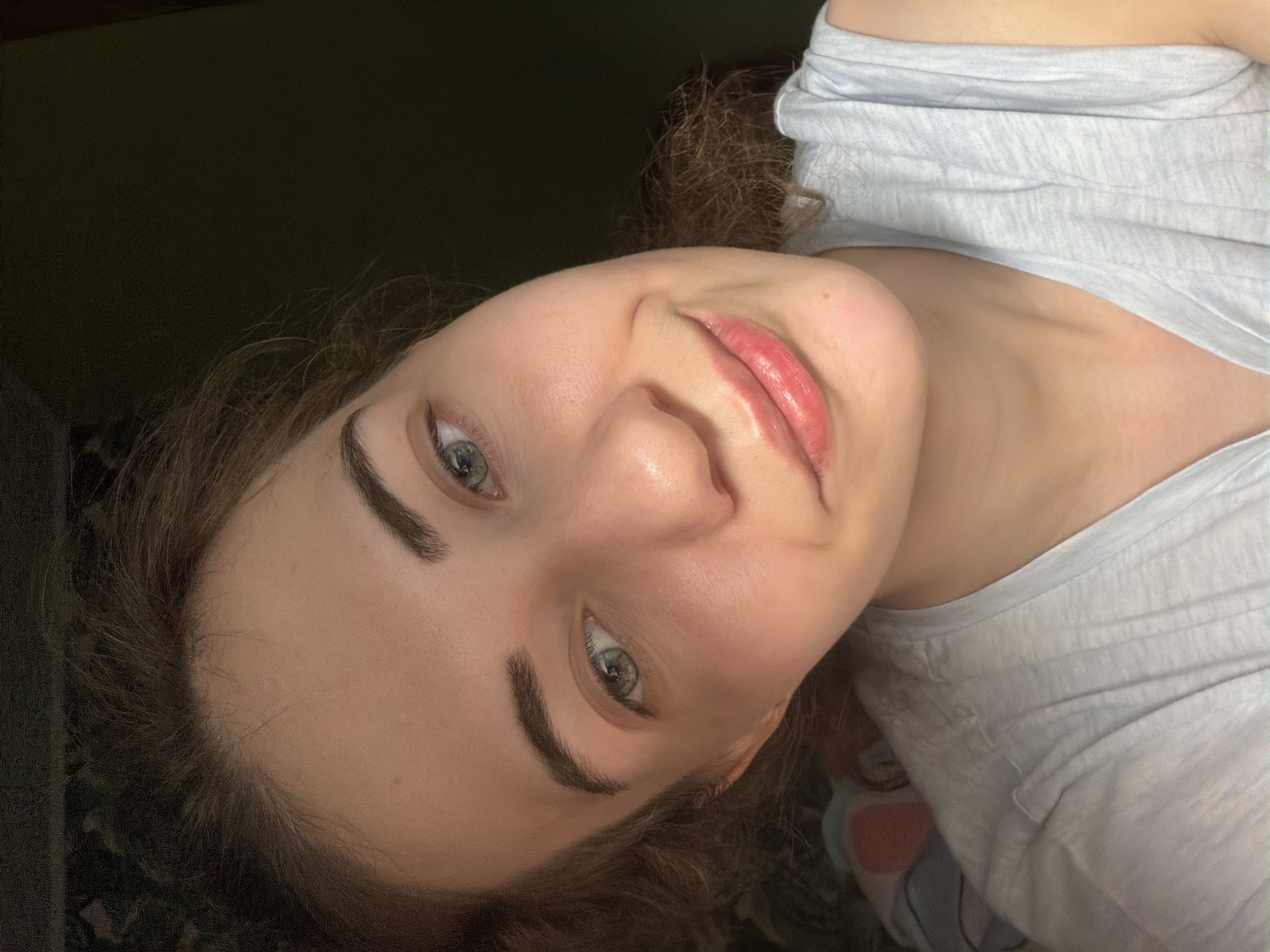 The image is a close-up selfie of a young woman with light skin, light brown curly hair, and light green eyes. She is wearing a light blue t-shirt. Her lips are slightly parted and glossy, and she has a subtle smile. The lighting suggests it is indoors, with a dark background. There is no information about the location of Zavodoukovsk, Russia in the image.
