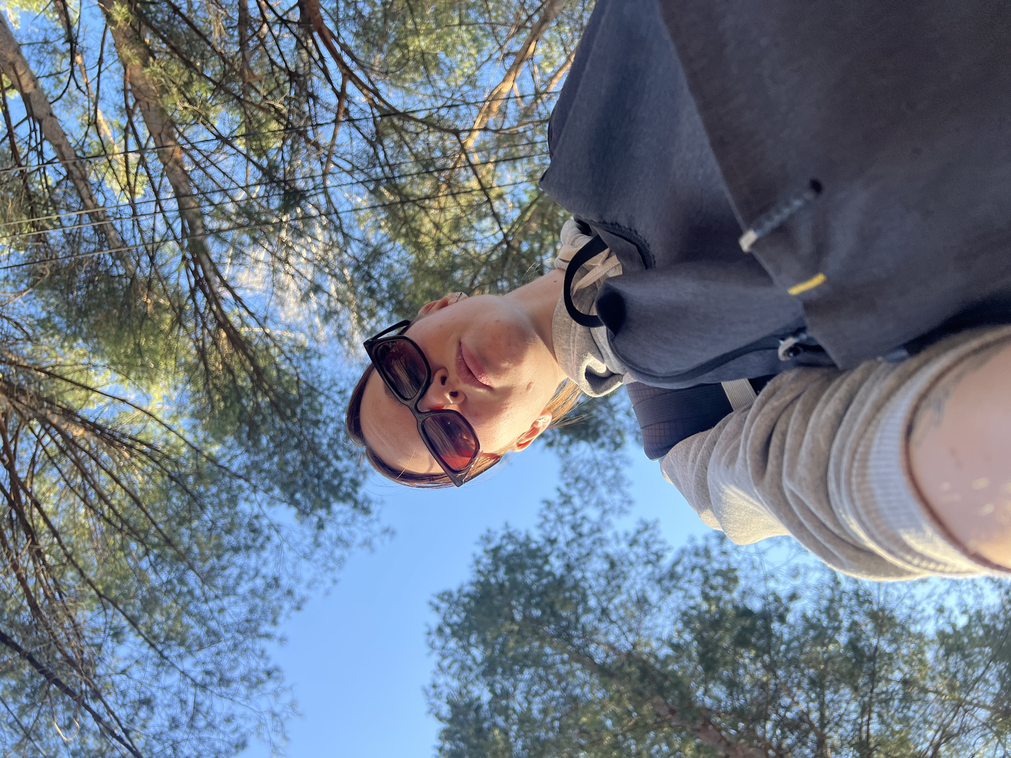 A low-angle shot depicts a woman wearing sunglasses. She is outside with trees and sky in the background. The location is not specified. The woman is wearing a gray and black hoodie. The sky is blue.