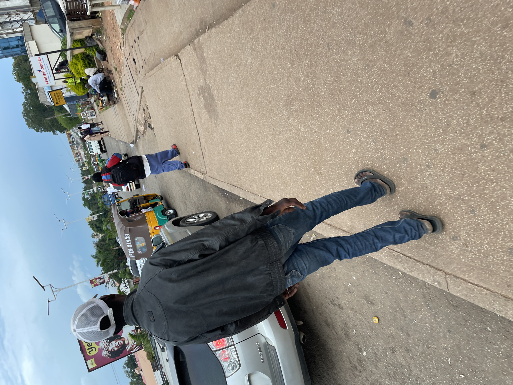 The image depicts a street scene in Jos, Nigeria. In the foreground, a person is walking away from the camera, wearing a black jacket, blue jeans, and a white mesh cap. They are on a concrete sidewalk next to a paved road. To the left, a silver SUV is parked. Further down the road, a colorful auto-rickshaw with the license plate "PTA 11478" is visible, along with other vehicles and pedestrians. Buildings line the street, including one with a sign for "XPRESSCOURIER". The sky is overcast.