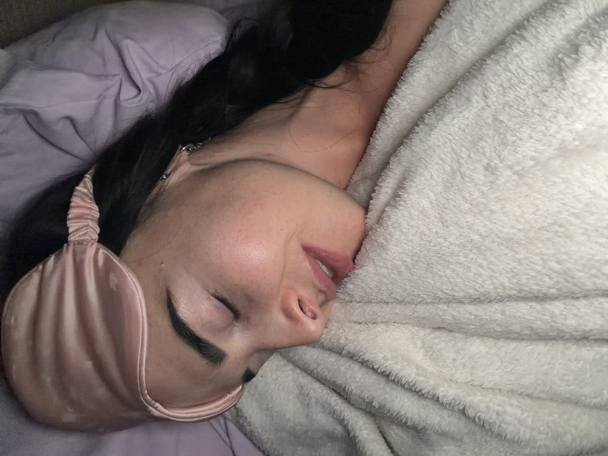 An overhead shot captures a person, likely female, lying on their side in what appears to be a bed, rotated clockwise. The individual has long, dark hair spread out on a light purple pillow or sheet. A pink, satin sleep mask covers their eyes, suggesting they are sleeping or deeply resting. Their facial features are visible from the nose down, with their mouth slightly ajar. The skin shows some freckles or natural skin texture.
The person is partially covered by a soft, white, fluffy blanket or towel, which is textured with ripples and folds, indicating a cozy environment. A small silver chain or piece of jewelry is faintly visible near their neck or earlobe. The lighting is soft and subdued, creating a peaceful ambiance. The overall scene suggests a quiet moment of sleep or relaxation indoors. No text is visible in the image.
