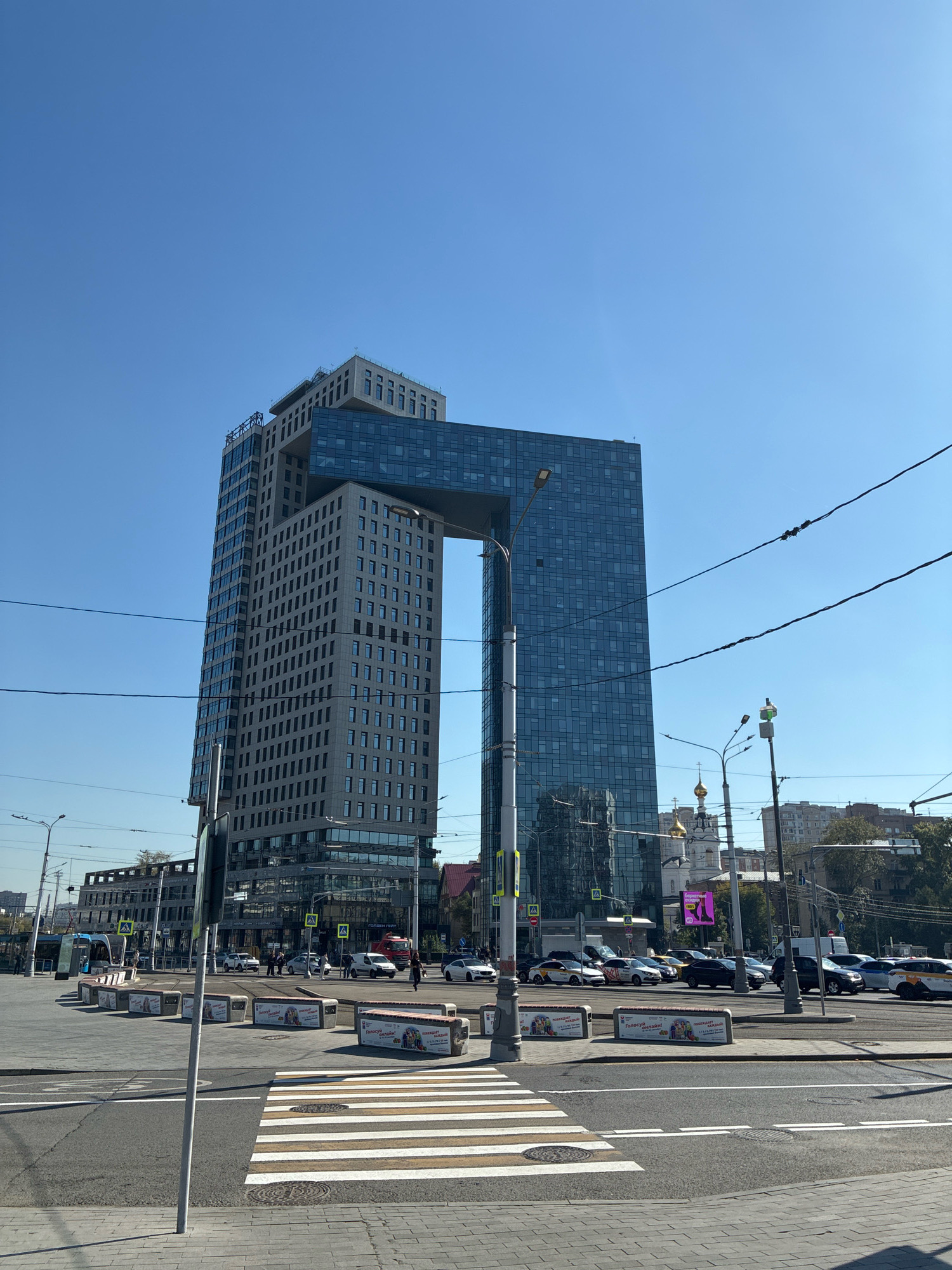 The image depicts the Moscow City business district in Moscow, Russia. The focal point is the modern, arch-shaped skyscraper known as "Dominion Tower," which features a distinctive gap in its structure. The skyscraper is a mixed-use building with offices and residential units. To the left of Dominion Tower is another tall building with a light-colored facade and numerous windows. The foreground shows a street scene with a crosswalk, traffic lights, overhead wires, and several cars on the road. In the background, a white church with golden domes is visible, along with other urban buildings and trees. The sky is clear and blue, indicating a sunny day. There are no people prominently featured in the image, but it can be inferred that it is a busy urban environment.