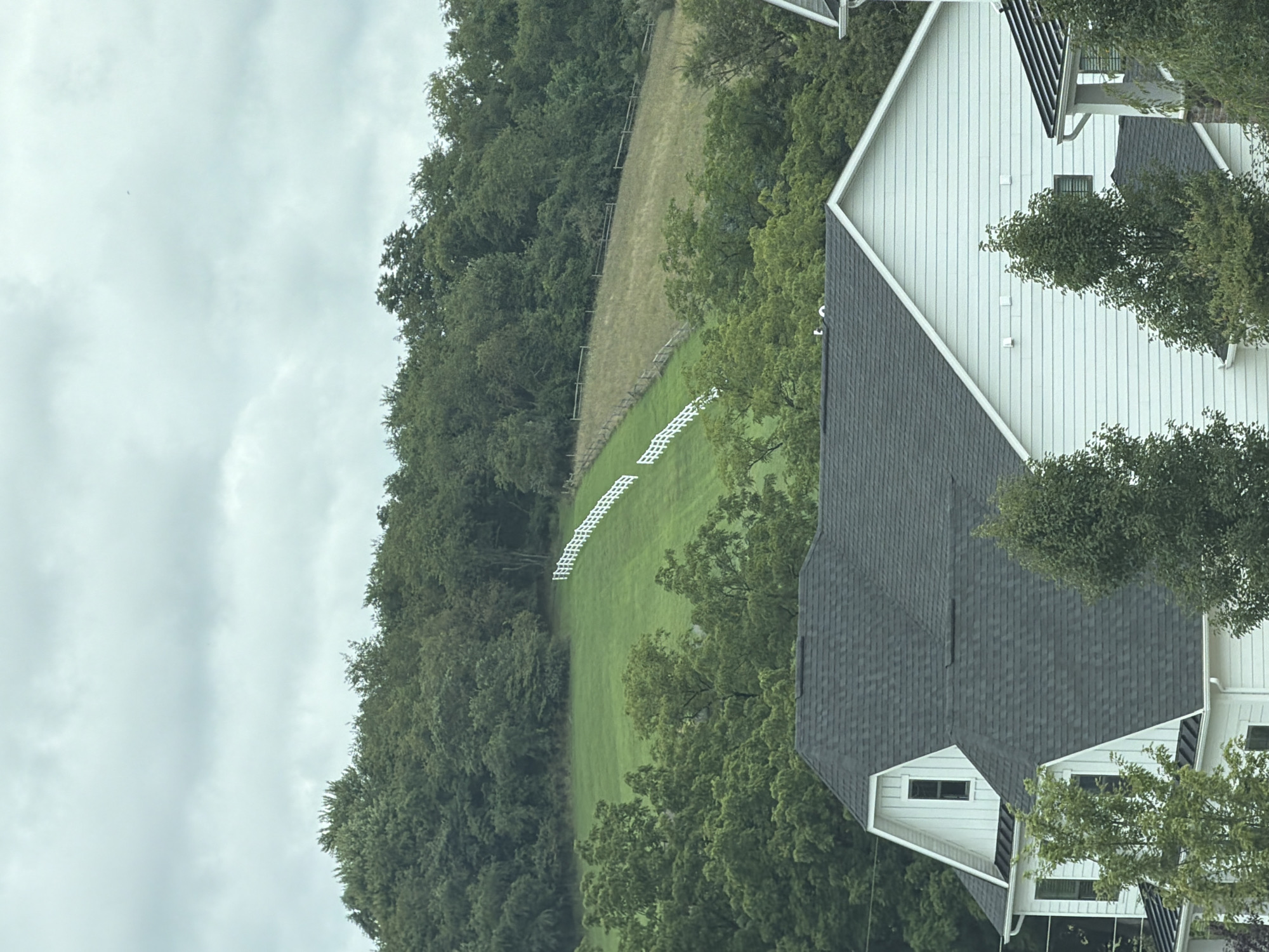 The image depicts a rural or suburban landscape with a focus on a green hillside. A white picket fence runs diagonally up the hill, demarcated by mown grass on one side and dry, brown grass on the other. The hillside is framed by dense green trees. In the foreground, the dark, shingled roof of a modern white house is visible, with architectural details like dormer windows and white siding. The sky is overcast with clouds. The scene is located in Wexford, United States of America.