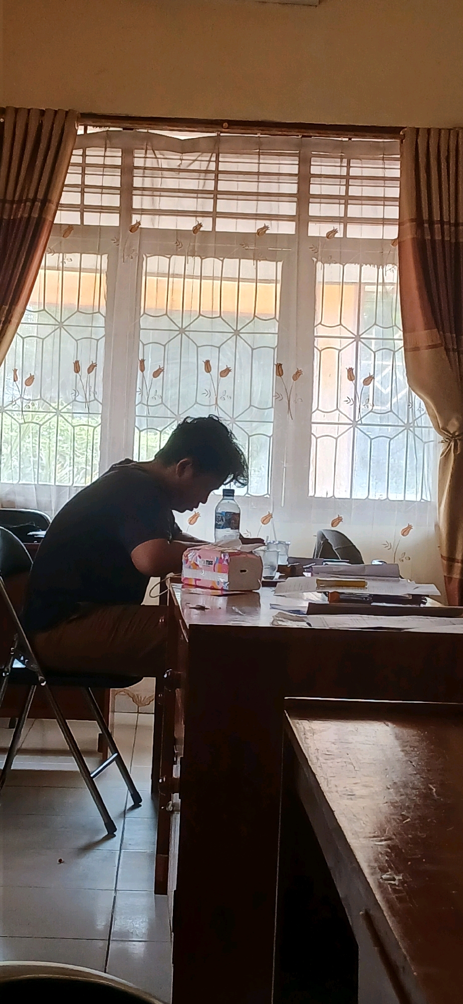 A person, identified as male based on physical attributes such as short hair and build, is seated at a wooden desk, engaged in an activity that appears to be studying or working. The individual is wearing a dark t-shirt and brown pants. In front of them, on the desk, is a box of tissues, a bottle of water, and various papers and writing implements. Behind the desk, a large window with a patterned curtain is visible, allowing natural light to enter the room. The flooring appears to be tiled. The setting is likely an indoor space, such as a classroom, office, or study room. The overall impression is one of focused activity within a functional environment. The image was taken in Metro, Indonesia.