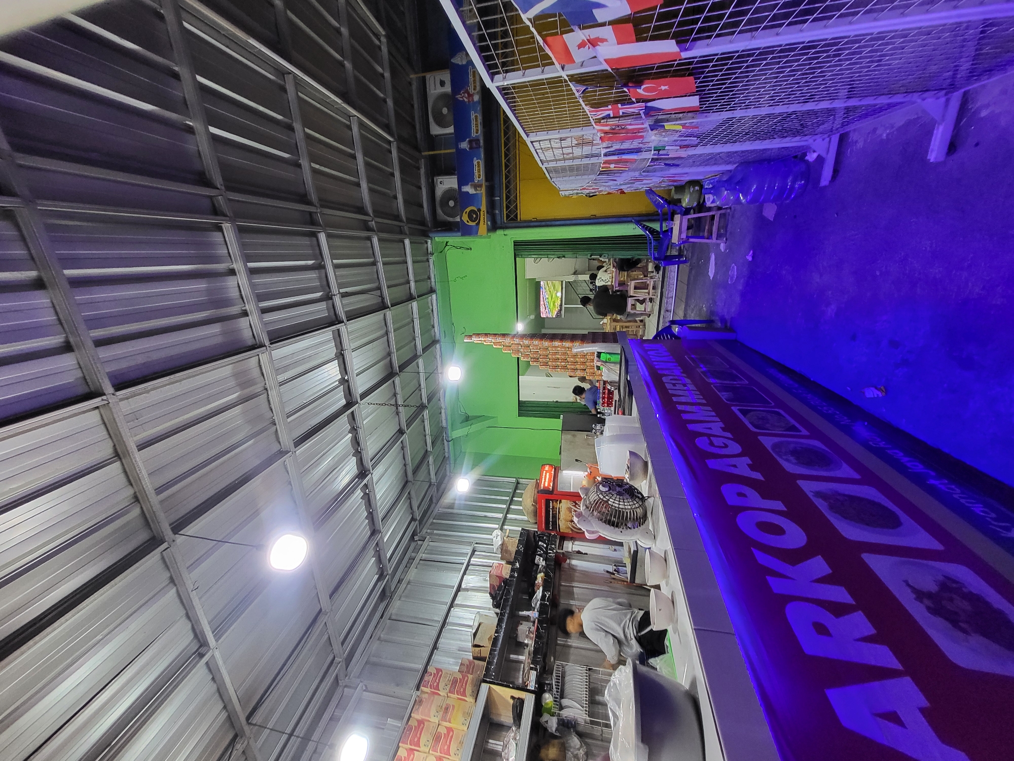 The image depicts the interior of a food stall in North Jakarta, Indonesia. The scene is characterized by a corrugated metal ceiling with exposed beams and fluorescent lighting. In the foreground, a long counter displays various bowls, a fan, and what appears to be food items or ingredients. A purple banner with white text and images runs along the front of the counter. The text on the banner is partially visible and reads "ARKOP AGAM MEDAN KO". Behind the counter, shelves are stocked with boxes, and a Coca-Cola refrigerator is visible. Further back, a brightly lit green wall serves as a backdrop. Open doorways reveal a seating area with tables and chairs, where several people are dining. A television screen is visible in one of the doorways, displaying a soccer match. To the right, a metal fence is decorated with a string of small national flags, including those of Canada, the United Kingdom, and Turkey. The overall impression is of a casual, bustling eatery.