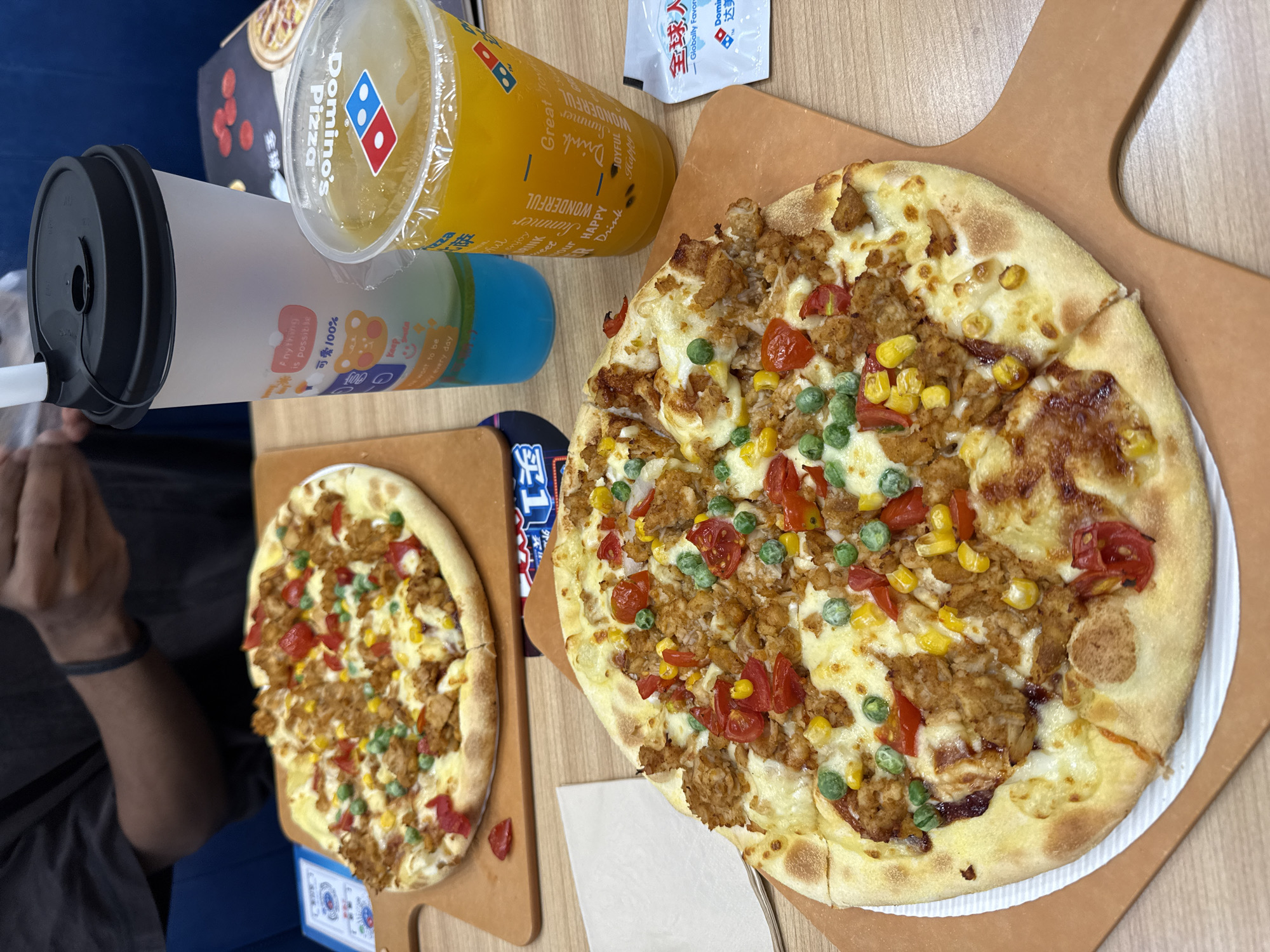 The image shows two pizzas on a wooden table at a Domino's Pizza restaurant in Dongcheng District, China. The pizza in the foreground is larger and has toppings of shredded chicken, corn, peas, and cherry tomatoes on a cheese base. The pizza in the background is smaller and appears to have similar toppings. Two drinks are visible: one is a blue beverage in a translucent cup with a black lid and straw, and the other is a yellow-orange beverage in a clear plastic cup with a Dominos logo. A small packet with Chinese writing and a Dominos logo is also present on the table. In the background, a person's arms are visible, wearing a dark t-shirt and a black wristband. The environment appears to be a casual dining setting.