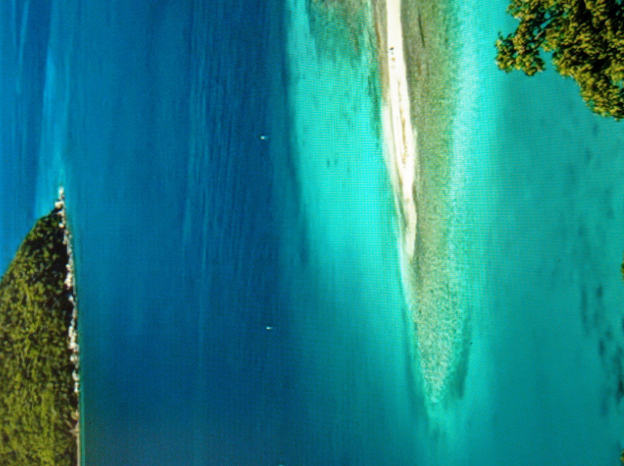 The image shows a tropical scene with lush green foliage on an island in the distance. The water is a vibrant turquoise, transitioning to a deep blue further out. A sandy bar or shallow reef is visible beneath the surface, creating a lighter, almost white area in the turquoise water. The image is a landscape photograph, possibly taken from a higher vantage point, showcasing the natural beauty of a coastal or island environment. There are no visible people or specific objects that would suggest a location within the city of Mukachevo, Ukraine, as this scene appears to be a tropical or subtropical seascape.