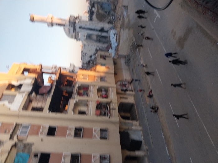 A street scene shows people walking on the asphalt road with buildings on the left and a mosque structure in the background. The building on the left appears to be an apartment complex, while the mosque has a minaret. The light suggests the photo was taken during the day. A cable is visible in the top right. The image is slightly rotated.