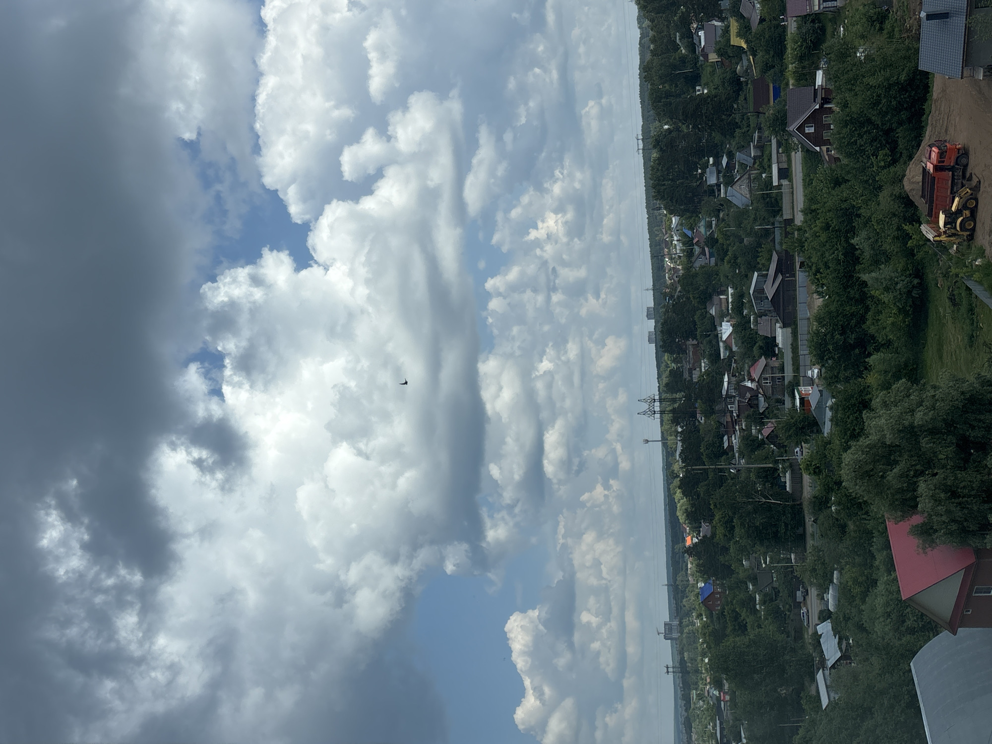 This aerial photograph depicts a suburban or rural landscape dominated by lush green trees and scattered residential buildings. The sky is filled with dramatic, large cumulus clouds, interspersed with patches of blue sky, suggesting dynamic weather conditions. A lone bird is visible in the upper portion of the frame, soaring against the clouds.
In the midground and foreground, numerous houses with various roof colors, including red, brown, and grey, are nestled amongst dense foliage. Some of these homes appear to be of a traditional dacha style, common in rural Russia. There are signs of construction or renovation, with a pile of sand and two construction vehicles—a yellow excavator and a red dump truck—situated in the lower right portion of the image. Power lines and transmission towers are also visible in the distance, indicating infrastructure present in the area. The overall impression is of a community integrated with its natural surroundings, with a hint of ongoing development.
While the specific location within the city of Chaikovsky, Russia, cannot be pinpointed definitively from the image alone, the architectural styles of the houses, the presence of dacha-like structures, and the general landscape are consistent with many settlements in the Perm Krai region. The scene appears to be captured during daylight hours, likely late morning or afternoon, given the lighting and cloud formations. There are no people directly visible in the photograph.