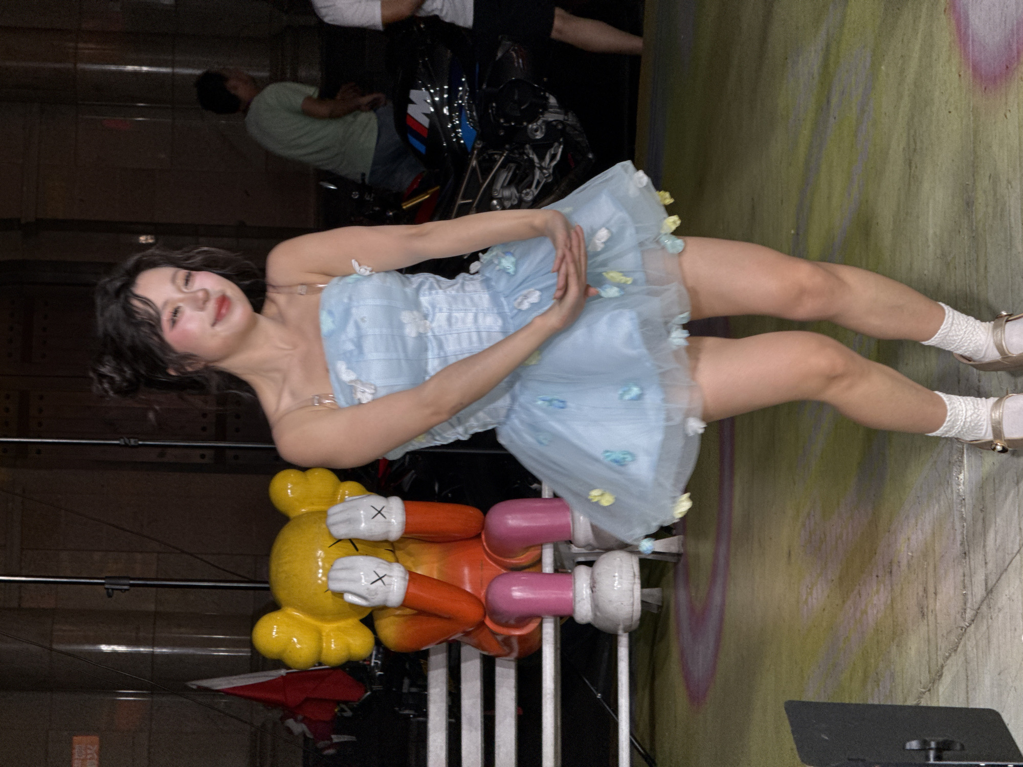 The image portrays a young woman posing in an indoor or covered urban-industrial setting, likely in Nan'an District, China. She lies on her side, facing the camera with a gentle smile and a soft expression. Her dark, wavy hair frames her face, which features prominent blush. She is dressed in a light blue strapless dress, characterized by a corseted bodice and a flowing, multi-layered skirt embellished with small, pastel-colored fabric flowers. Her outfit is completed with white crocheted socks and light-colored mary-jane style shoes.
To her left, on the textured concrete floor, is a large, colorful KAWS Companion figure, featuring a yellow head, orange arms, and pink legs, with white hands and feet marked by black "X"s. The concrete floor exhibits faded graffiti or paint in various shades of green, yellow, and pink. In the darker background, another person in a green shirt and jeans is partially visible, along with parts of a motorcycle or scooter and metal scaffolding. A black rectangular object, possibly equipment, is seen in the bottom right corner. The scene suggests a photoshoot or video production due to the woman's posed posture and the artificial lighting conditions. No discernible text is visible within the frame.