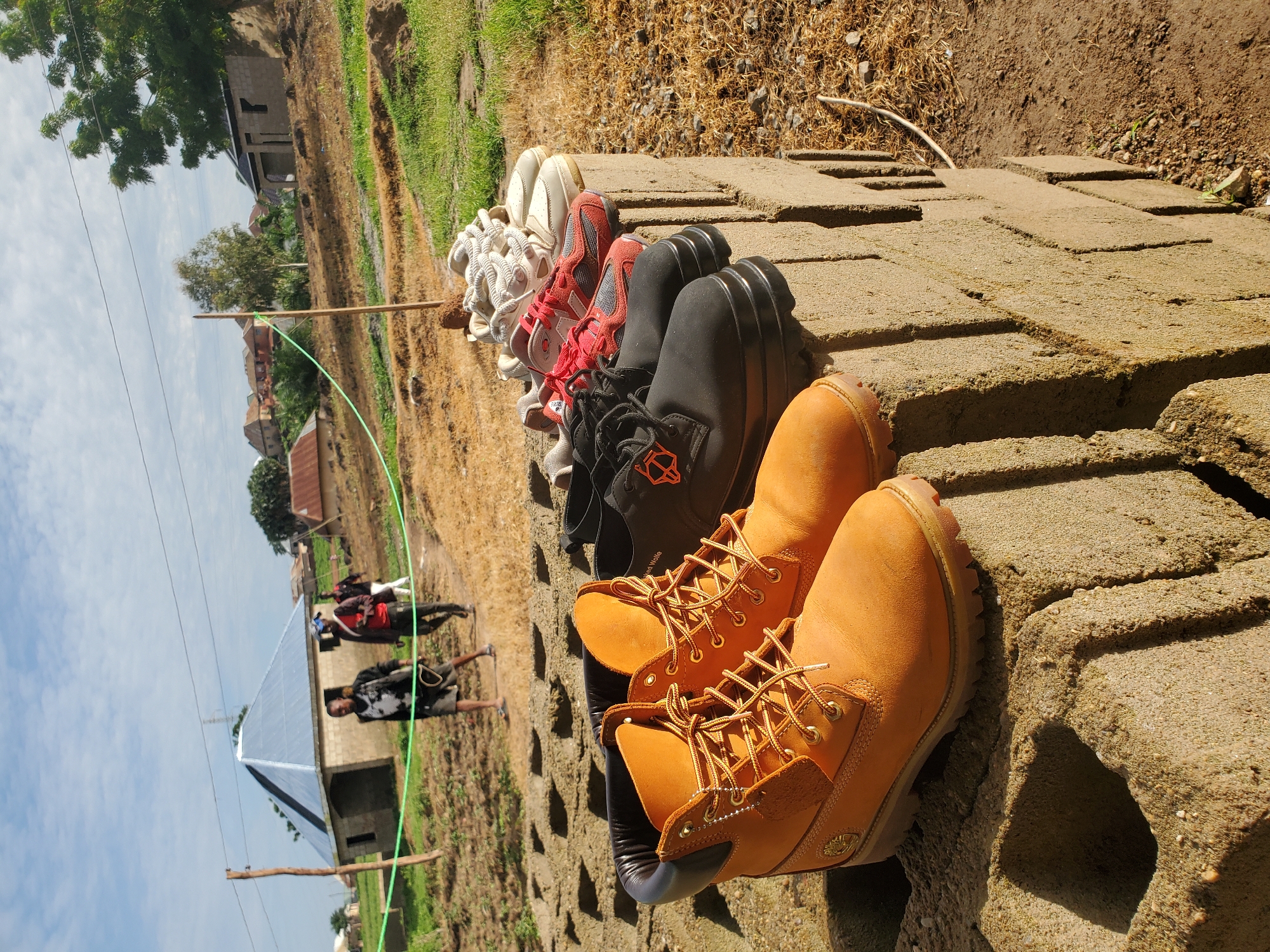 The scene is set in the city of Winters, United States of America, under a bright, partly cloudy sky. In the foreground, a stack of rough, light brown concrete blocks serves as a display surface for five pairs of shoes lined up horizontally. From left to right, these include a prominent pair of tan work boots with yellow laces, a pair of black lace-up shoes with thick soles and an orange geometric logo, a pair of red and grey athletic shoes, an off-white pair of athletic shoes with chunky soles, and a partially visible pair of white athletic shoes. The immediate ground is dry, reddish-brown soil mixed with sparse dry grass and small stones.
In the midground, a dirt path stretches diagonally, occupied by three individuals walking away from the viewer. The foremost person wears dark shorts and a patterned top, appearing to walk barefoot or in sandals. Behind them, a second person wears dark trousers and a red shirt, carrying a white object over their shoulder. The third individual is further back, dressed in dark attire. A light green rope or hose is suspended across the midground, supported by at least two wooden poles.
The background features a collection of buildings and natural elements. On the left, a single-story structure with light-colored concrete block walls and a distinctive shiny grey pyramidal roof is visible. Further right, other simple dwellings and a partially constructed building with exposed concrete block walls are present. Scattered trees, some with green foliage, punctuate the landscape of dry earth and sparse vegetation. Overhead, power lines stretch across the bright sky, which is dotted with scattered white clouds, indicating clear daytime conditions.