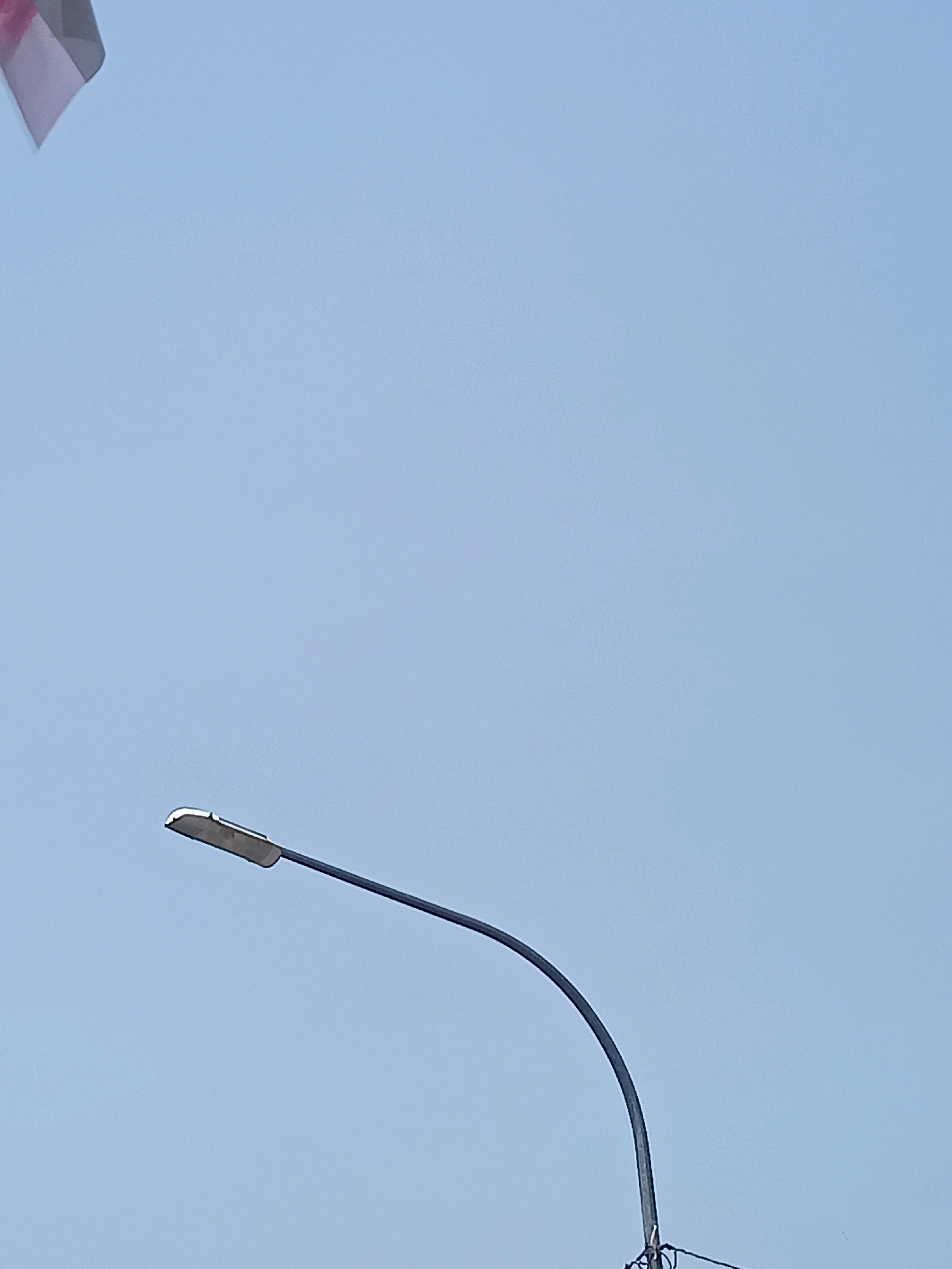 The image shows a street lamp against a clear, light blue sky. In the upper left corner, a small portion of a flag is visible, appearing to be red and white. The street lamp's arm curves from the bottom right towards the top left, with the lamp fixture itself positioned in the lower half of the frame. No people or other objects are discernible in the image. The scene is an outdoor environment, likely a public space in Gabungan, Indonesia, characterized by the presence of street lighting infrastructure.