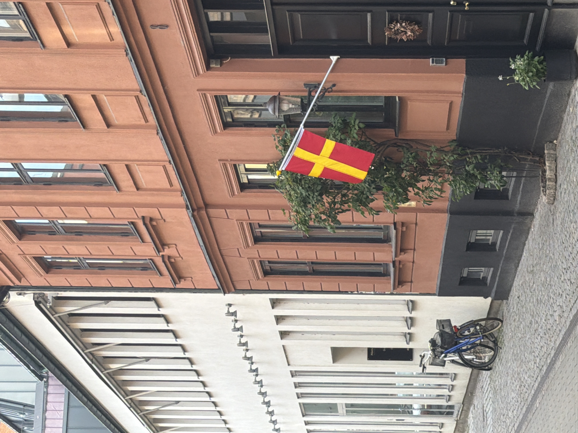 The image shows a street scene in Malmö, Sweden. On the right is a building with an orange-brown facade, featuring a black door with a decorative wreath and a digital lock. A Danish flag hangs from a pole attached to the building, alongside a vintage-style lamp. A small tree with green foliage grows in front of the building, partially obscuring the lower windows. To the left of the orange-brown building is a white building with a modern design, characterized by numerous vertical windows and a row of exterior lights. Two bicycles, one blue and one lighter colored, are parked on the cobblestone street in front of the white building. The street is paved with cobblestones, and a raised sidewalk is visible. The overall scene depicts an urban environment with a mix of architectural styles.