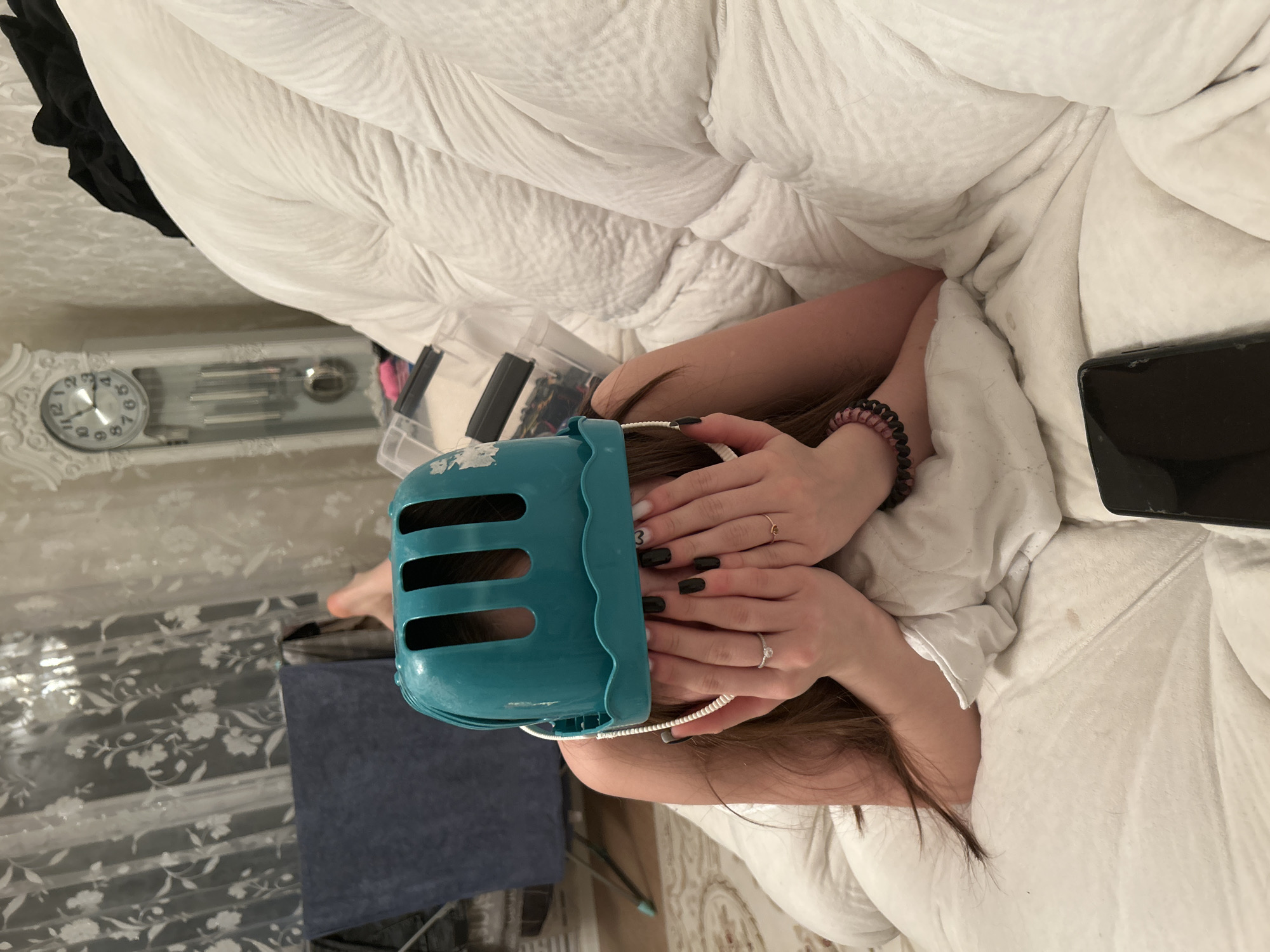 This image depicts an indoor scene within a residential setting in Chaikovsky, Russia. A person, identified by long brown hair, is lying prone on a white quilted bedspread, facing away from the camera's direct line of sight. They are covering their face with both hands. A turquoise plastic colander, with vertical slots and a wavy rim, is worn on their head, resembling a helmet. A white cord is visible beneath the colander on the left side of their head.
Essential details about the person include:
* Fingernails painted black, with one fingernail on the right hand featuring a white design, possibly a heart.
* A silver-colored ring with a clear stone on the ring finger of the left hand.
* A thin gold-colored ring on the ring finger of the right hand.
* A pink and black coiled hair tie bracelet on the right wrist.
The immediate foreground to the lower right shows a black smartphone with a visible crack on its screen, resting on the white bedding.
The background elements include:
* A white grandfather clock with a decorative frame, a round face displaying time close to 1:55, and three pendulums, positioned to the upper right behind the person.
* Two clear plastic storage containers with black lids stacked near the clock.
* White sheer curtains with a floral pattern covering a window on the left.
* A dark blue or grey towel or fabric draped over a drying rack or piece of furniture in the mid-left background.
* Light-colored wallpaper with a subtle pattern is visible on the wall.
* A patterned rug or carpet is partially visible on the floor to the lower left.
The lighting suggests an indoor artificial source. The scene appears to be a casual moment within a bedroom or living area.