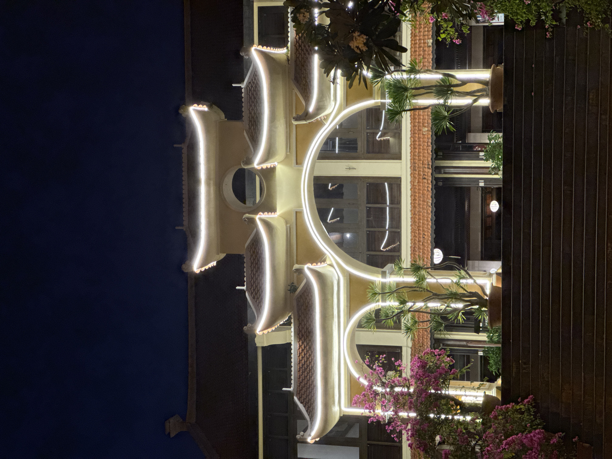 The photograph shows a multi-story building in Da Nang, Vietnam at night. The building is illuminated with decorative white lights outlining its architectural features. It appears to be a commercial property. The sky is dark blue and the building has a tiled roof. There are potted plants decorating the facade, including trees with green leaves and pink flowers. A dark brown wooden fence is visible on the right side.